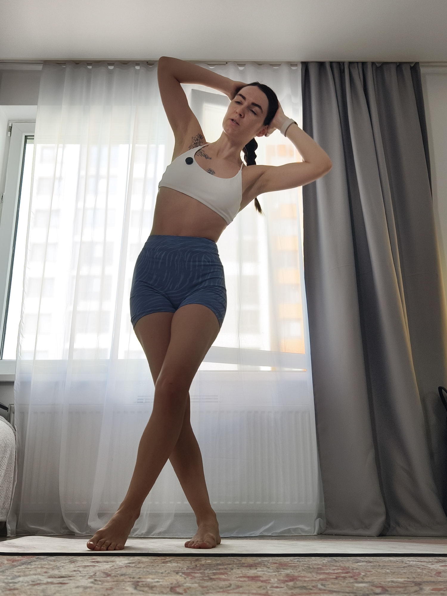 A full-body, low-angle shot captures a woman performing a stretching or yoga pose indoors. She stands in the center of the frame, facing slightly right, with her torso angled towards the left. Her arms are bent, with hands interlocked behind her head and neck, and her body is gently tilted to the left. Her legs are crossed at the ankles, with the right leg in front of the left. She wears a white sports bra and blue high-waisted shorts with a subtle wave pattern. A white wristband is visible on her right wrist, and she has a floral-like tattoo on her left bicep. Her eyes are closed, conveying a sense of focus and calm.
The setting appears to be a home in Tyumen, Russia, likely an apartment. Bright natural light streams from a large window directly behind her, which is covered by sheer white curtains. Through the curtains, distant multi-story buildings are faintly visible. To the right of the window, grey blackout curtains are drawn. A white radiator is visible below the window on the left. The woman stands on a light-colored yoga mat placed over a patterned rug or carpet. The ceiling is plain white. The bright lighting suggests it is daytime.