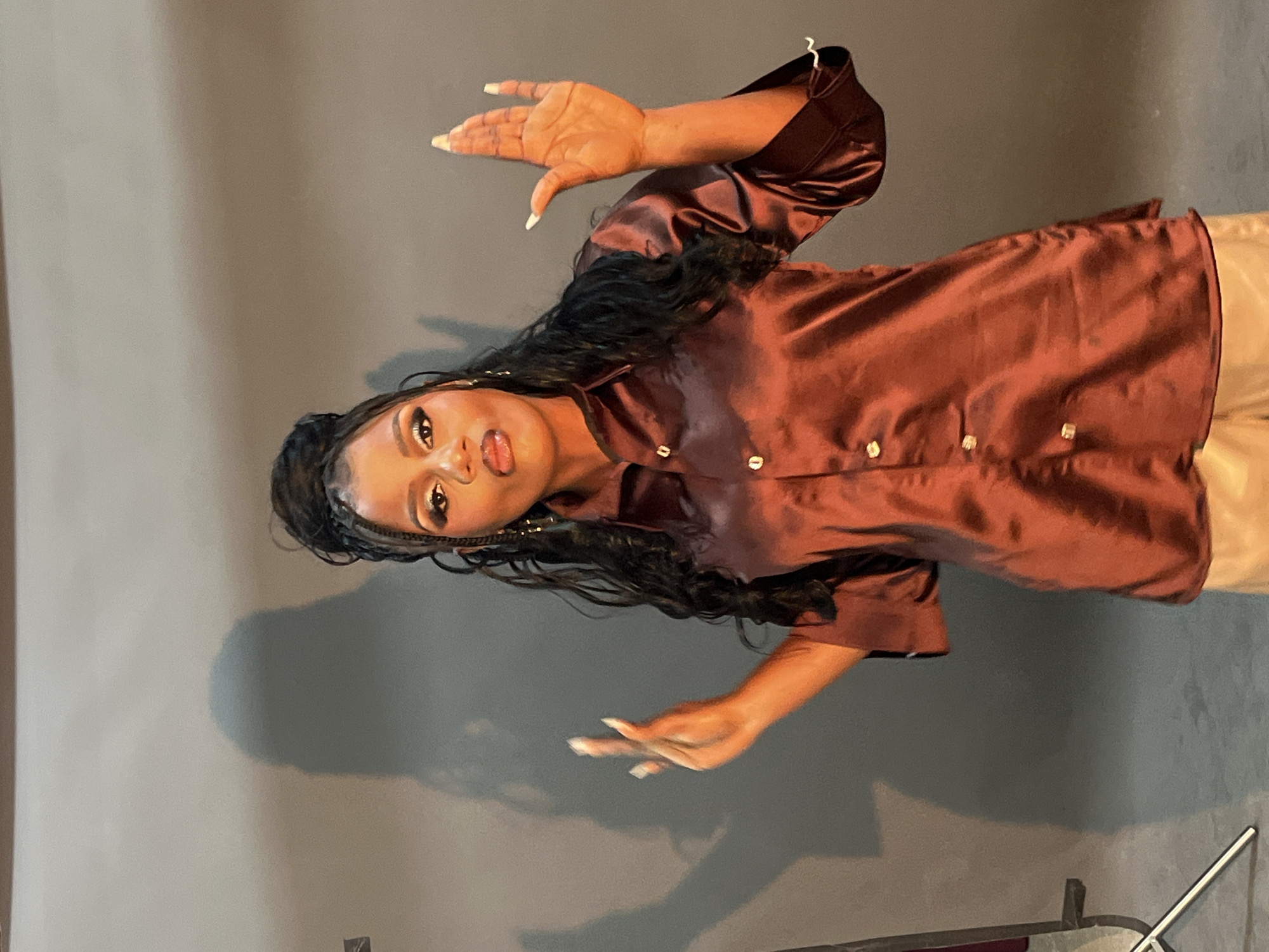 A woman with dark skin, long dark hair styled in a high bun with braids, and dramatic eye makeup stands against a plain gray studio backdrop. She is wearing a shiny, dark red button-up shirt with the sleeves rolled up and light-colored pants. Her hands are raised, with her left hand positioned in front of her chest with fingers slightly splayed, and her right hand held up with fingers extended and spread apart. A shadow of the woman is cast on the wall behind her. The setting appears to be a professional photo studio. No specific location in Gura Top, Nigeria, can be identified from the image.