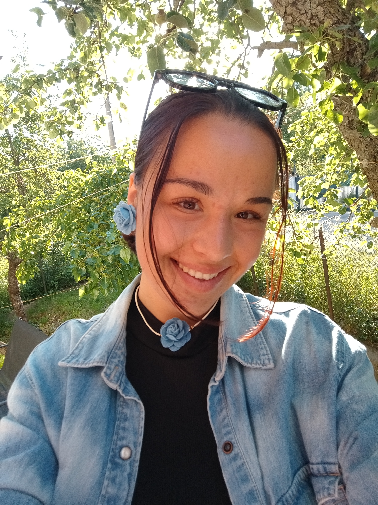 A selfie taken in an outdoor location in the city of Unknown, Unknown, shows a smiling woman with brown hair pulled back and sunglasses perched on her head. She is wearing a black turtleneck sweater and a light blue denim jacket. She is accessorized with light blue floral earrings and a matching necklace. The background features greenery, including trees and possibly a garden. Sunlight is filtering through the leaves, creating a dappled lighting effect. A wire fence is also visible in the background.