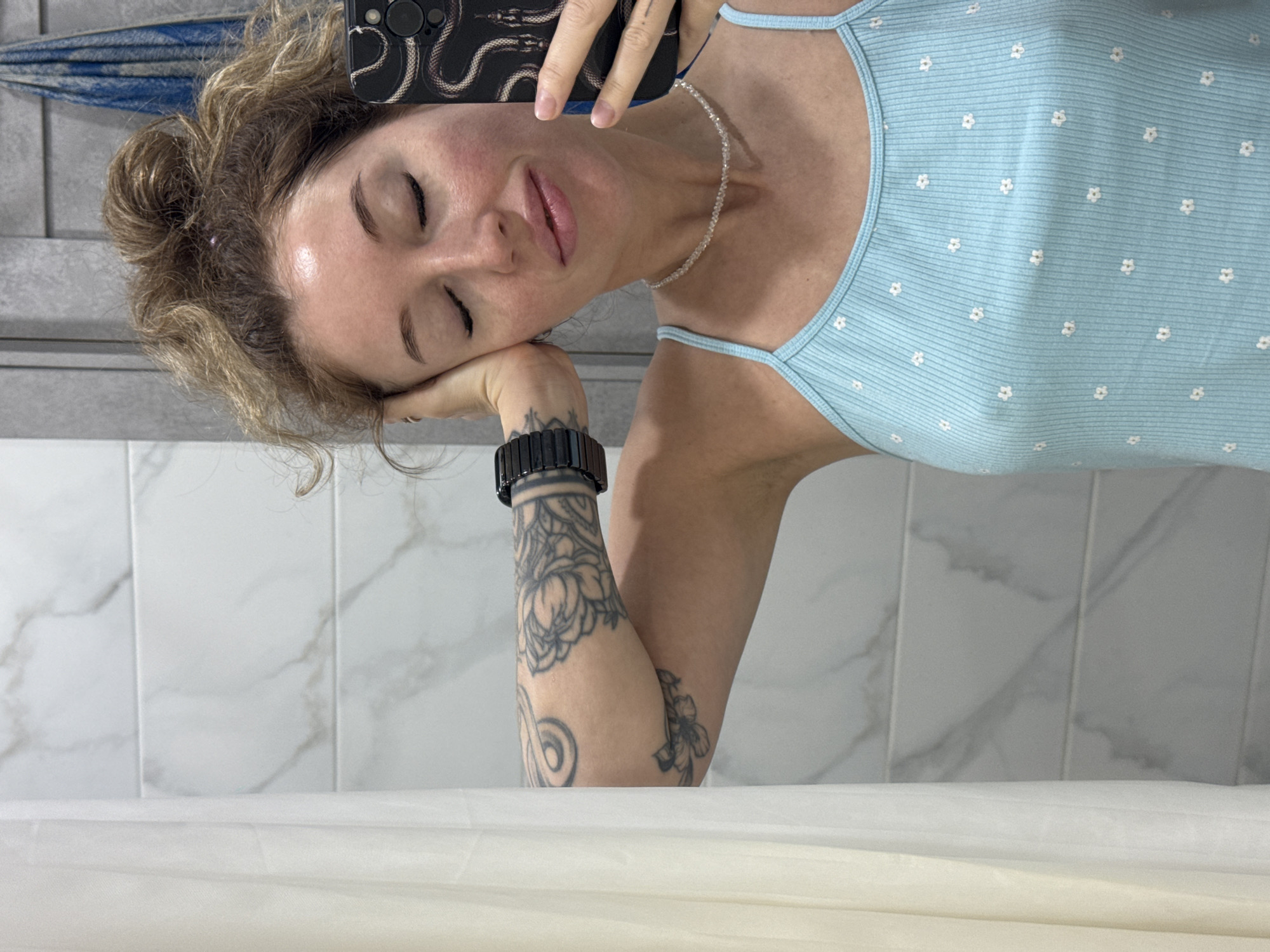 A young woman with curly blonde hair pulled up in a messy bun takes a selfie in a bathroom. She is wearing a light blue ribbed tank top with a floral pattern and a beaded necklace. Her right arm is raised, supporting her head as she closes her eyes. She has tattoos on her left arm and hand. The background shows a tiled wall, a shower curtain, and a towel hanging on a rack. The image appears to be taken indoors, possibly in a residential bathroom. The city of Chaikovsky, Russia, is known for its residential areas and public amenities, which would likely include bathrooms similar to the one depicted.
