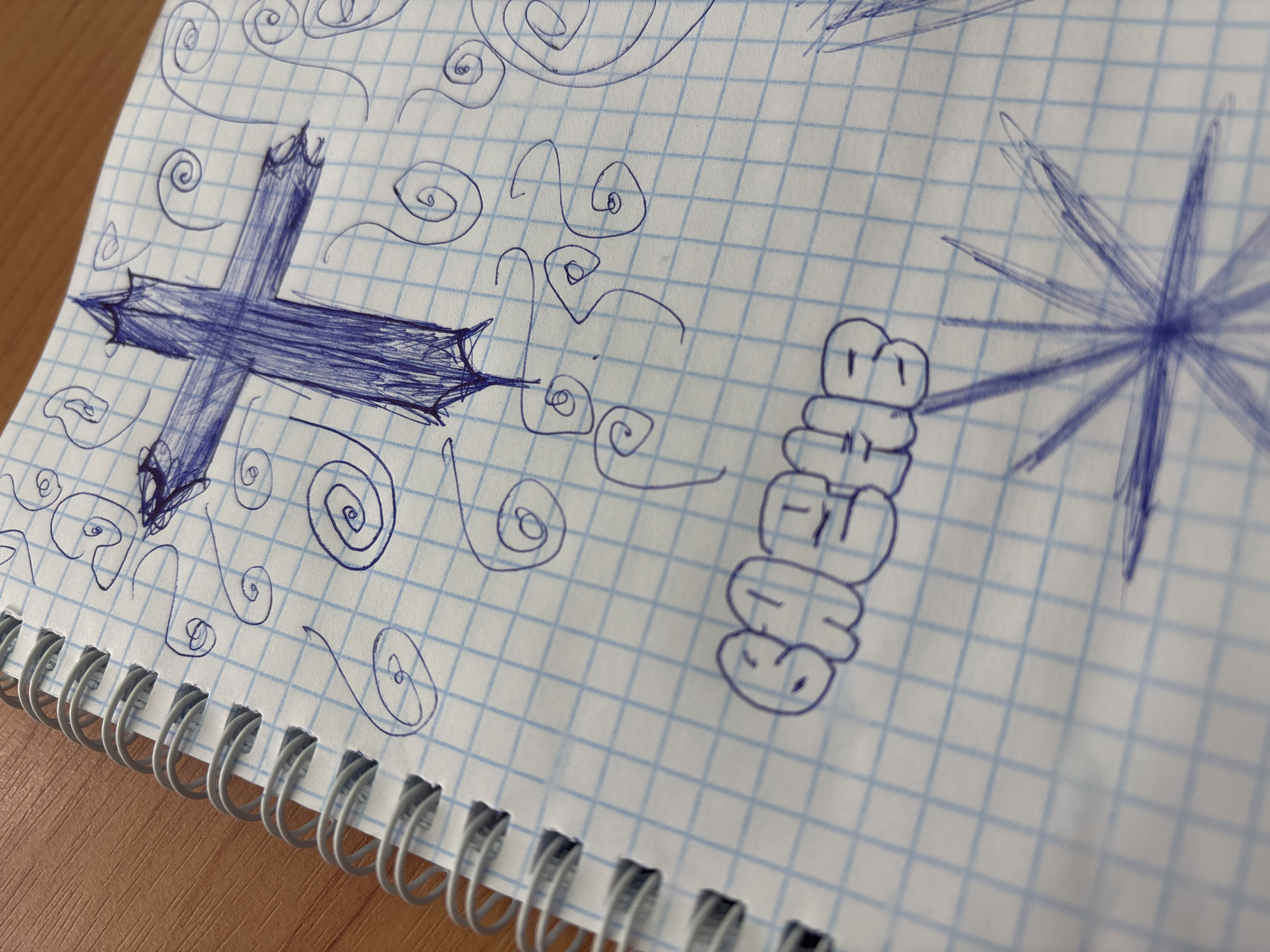 An overhead shot captures a page from a spiral-bound notebook, filled with various doodles drawn in blue ink. The paper is white with a faint light blue grid pattern. The notebook's silver metallic spiral binding is visible along the left edge of the page.
On the left side of the page, a prominent, roughly drawn blue cross is depicted. Its arms and top feature jagged, pointed ends, giving it a somewhat stylized or aggressive appearance. Surrounding this cross and scattered across the page are numerous small, intricate spirals and wavy, swirling lines, all drawn in the same blue ink.
Towards the center-right, a distinctive cartoonish drawing appears. It consists of five stacked, plump, rounded shapes. The top shape has two small dots resembling eyes. The shapes appear to form letters, possibly spelling "EHEHE" vertically, with each shape representing a letter.
Further to the right, another abstract design is present, resembling a starburst or a spiky, radiating circle, also drawn in blue ink with multiple lines emanating from a central point. The background surface, visible at the top and bottom left, appears to be a light wooden table or desk. The overall impression is a collection of casual, abstract sketches or doodles on graph paper. No specific time of day or location is discernible beyond an indoor setting.