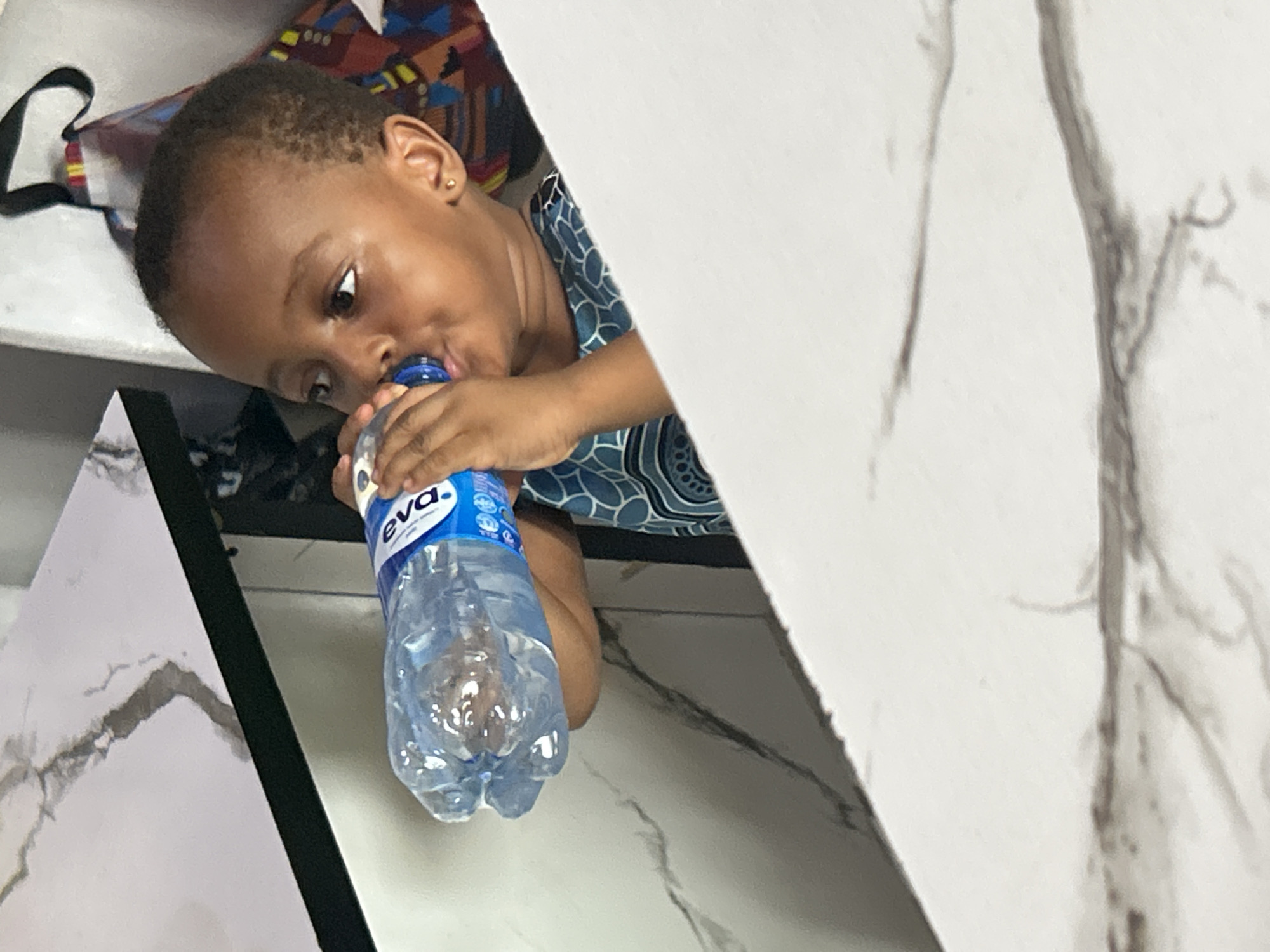 A young child is drinking from a bottle of "eva." water. The child is lying on a surface near a table with a marble pattern. The location appears to be a domestic interior, possibly in Ihiagwa, Nigeria, based on the brand of bottled water. The child is wearing a blue patterned shirt.
