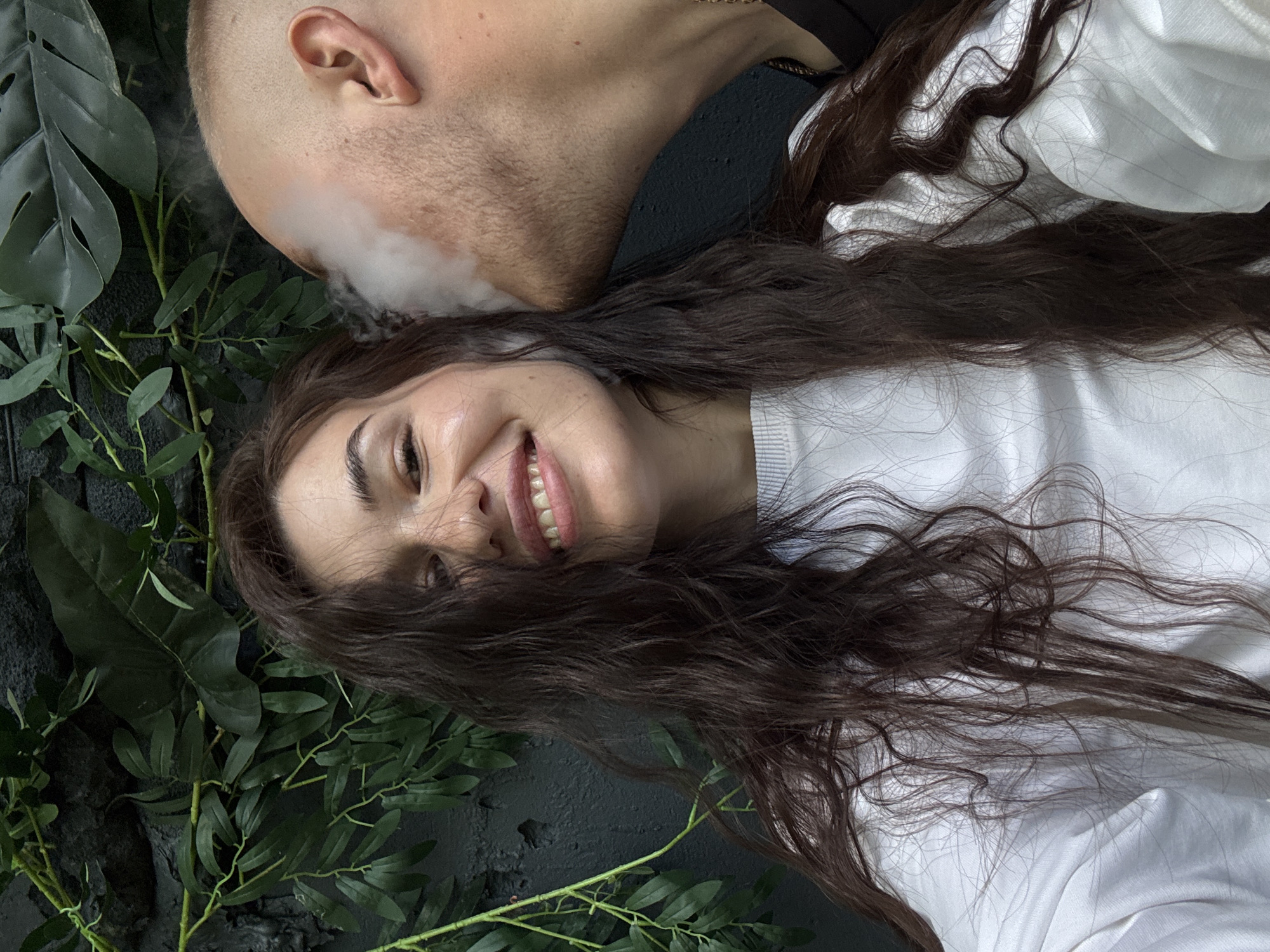 A close-up, selfie-style photograph captures a woman and a man in what appears to be an indoor setting with a dark, textured wall and green foliage as a backdrop. The woman is in the foreground, facing the camera with a wide smile, her long, dark, wavy hair framing her face. She is wearing a white, long-sleeved top. A man's head and neck are visible to her right, with smoke emanating from his mouth towards her ear. He has a shaved head and is wearing a dark shirt and a gold chain. The lighting is warm and direct, highlighting the subjects and the smoke. The presence of foliage and the dark wall suggest an urban environment, possibly a trendy establishment in Chaikovsky, Russia, though no specific landmarks are visible. The image focuses on the interaction between the two individuals, capturing a moment of shared activity.