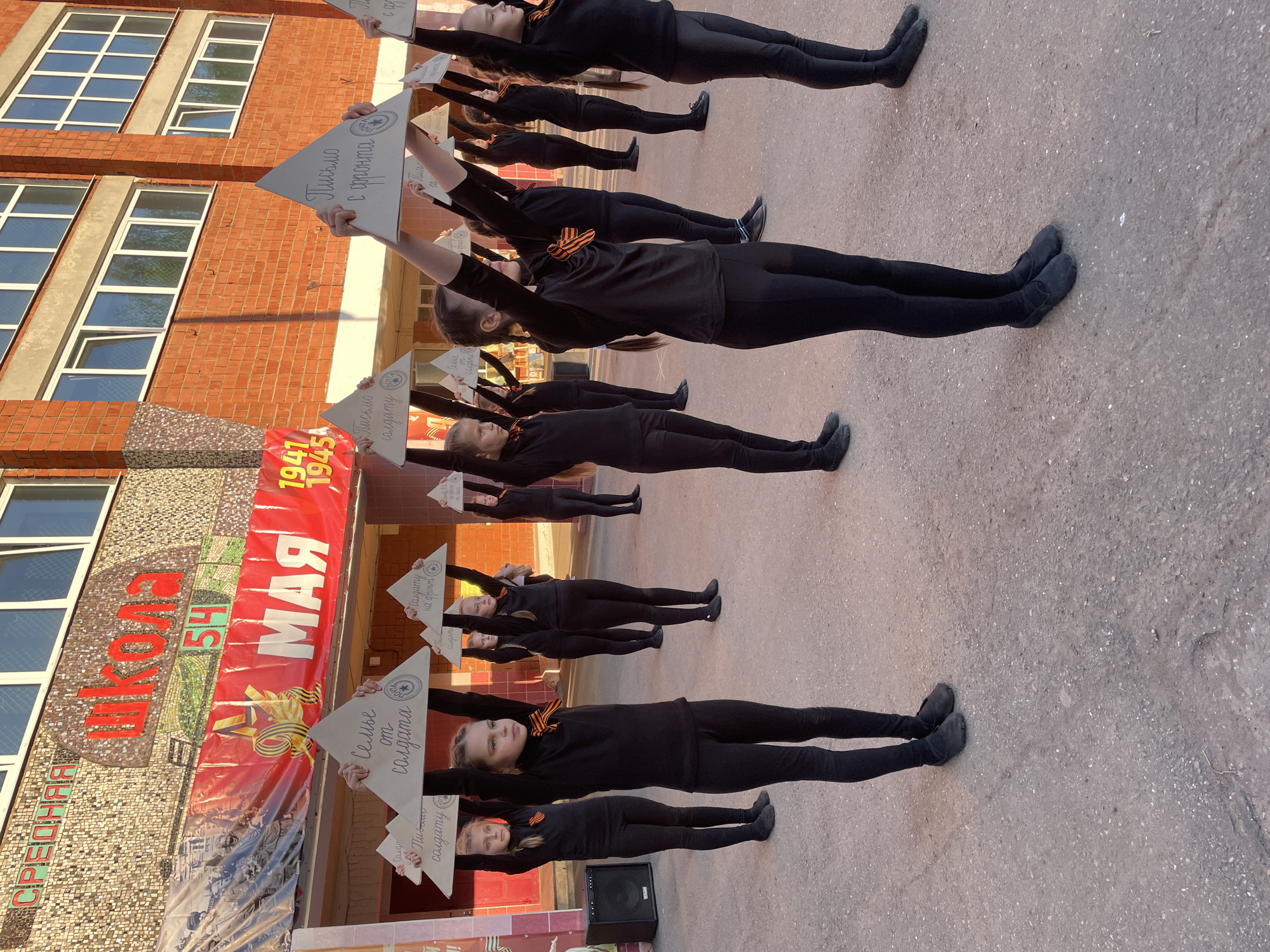 A group of at least twelve children, presumably students, are arranged in rows on what appears to be a gray, asphalt surface. They are all wearing black clothing and black shoes. Each child holds a triangular white sign above their head. The text on the signs appears to be written in Russian.
The background features a brick building with windows. A banner is displayed on the building with the word "МАЯ" (May) and the years "1941-1945," suggesting a celebration or commemoration related to World War II. A second banner with the word "ШКОЛА" (School) is also displayed on the left side of the building.
The overall scene indicates a public event, potentially a school assembly or commemorative activity. The location, based on text in the picture, is likely Izhevsk, Russia.