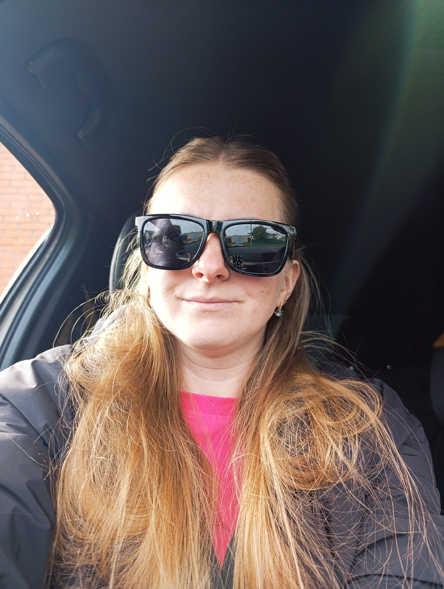 A woman is in a car, wearing sunglasses, a black jacket, and a pink shirt. She has long, reddish-blonde hair. The car appears to be parked on a street in a residential area. This is based on the reflection in the sunglasses, which shows houses and a street.