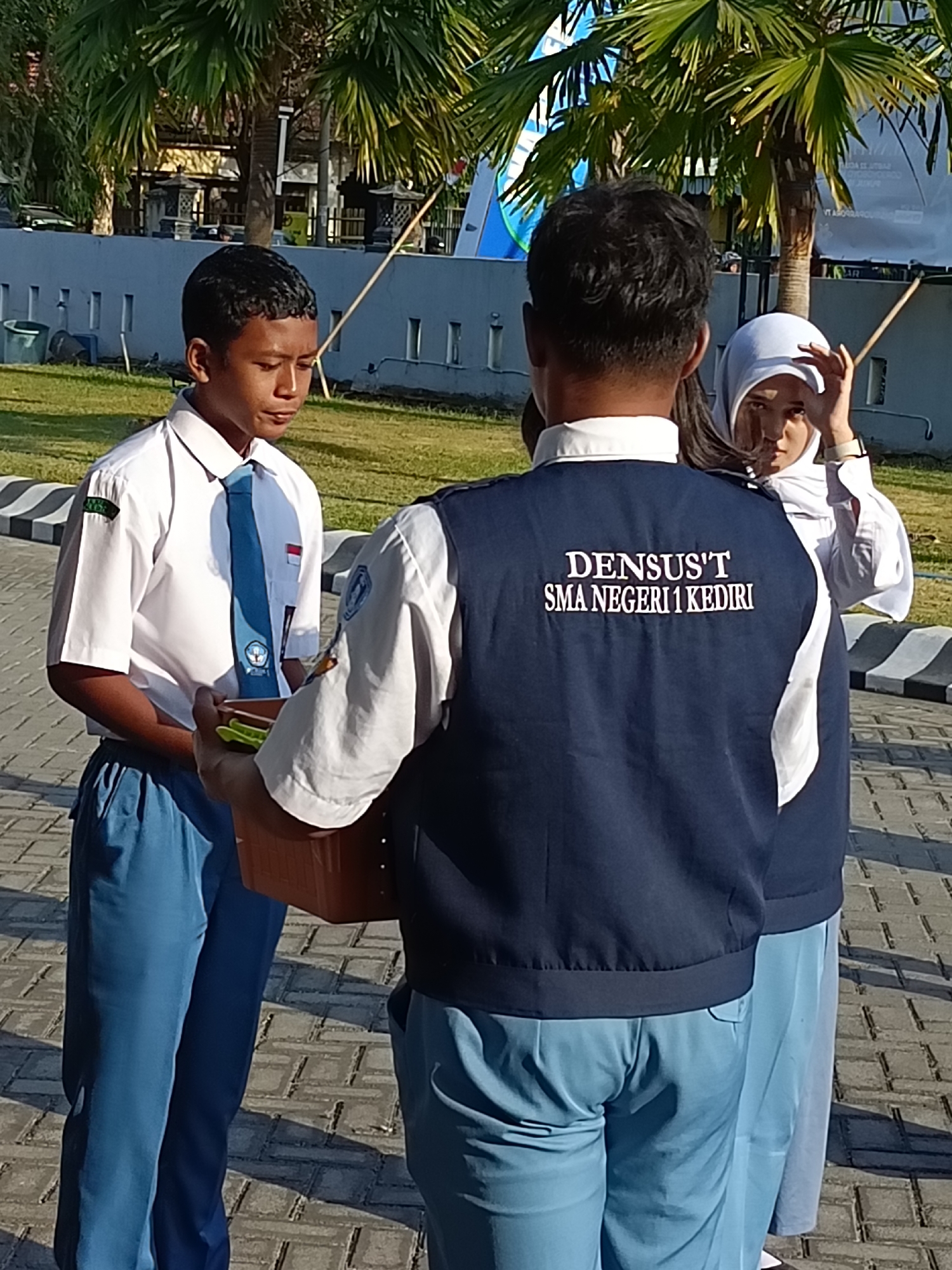 Three young people, likely students in school uniforms, are interacting outdoors. The individual on the left, a male, wears a white short-sleeved shirt, a blue tie with a crest, and blue trousers. He holds a brown rectangular object, possibly a box or container, in his hands. In the center, another individual, also male, is seen from the back. He wears a dark blue vest over a white shirt and blue trousers. The vest has white lettering that reads "DENSUS'T SMA NEGERI 1 KEDIRI". The individual on the right, a female, wears a white hijab and a white long-sleeved shirt, along with blue trousers. She is looking towards the camera and touching her hijab with her right hand. The background shows palm trees, a light blue wall, and a paved area with a patterned walkway in the foreground. The scene appears to be set in a sunny outdoor environment, possibly a school campus in Kediri City, Indonesia, given the text on the vest.
