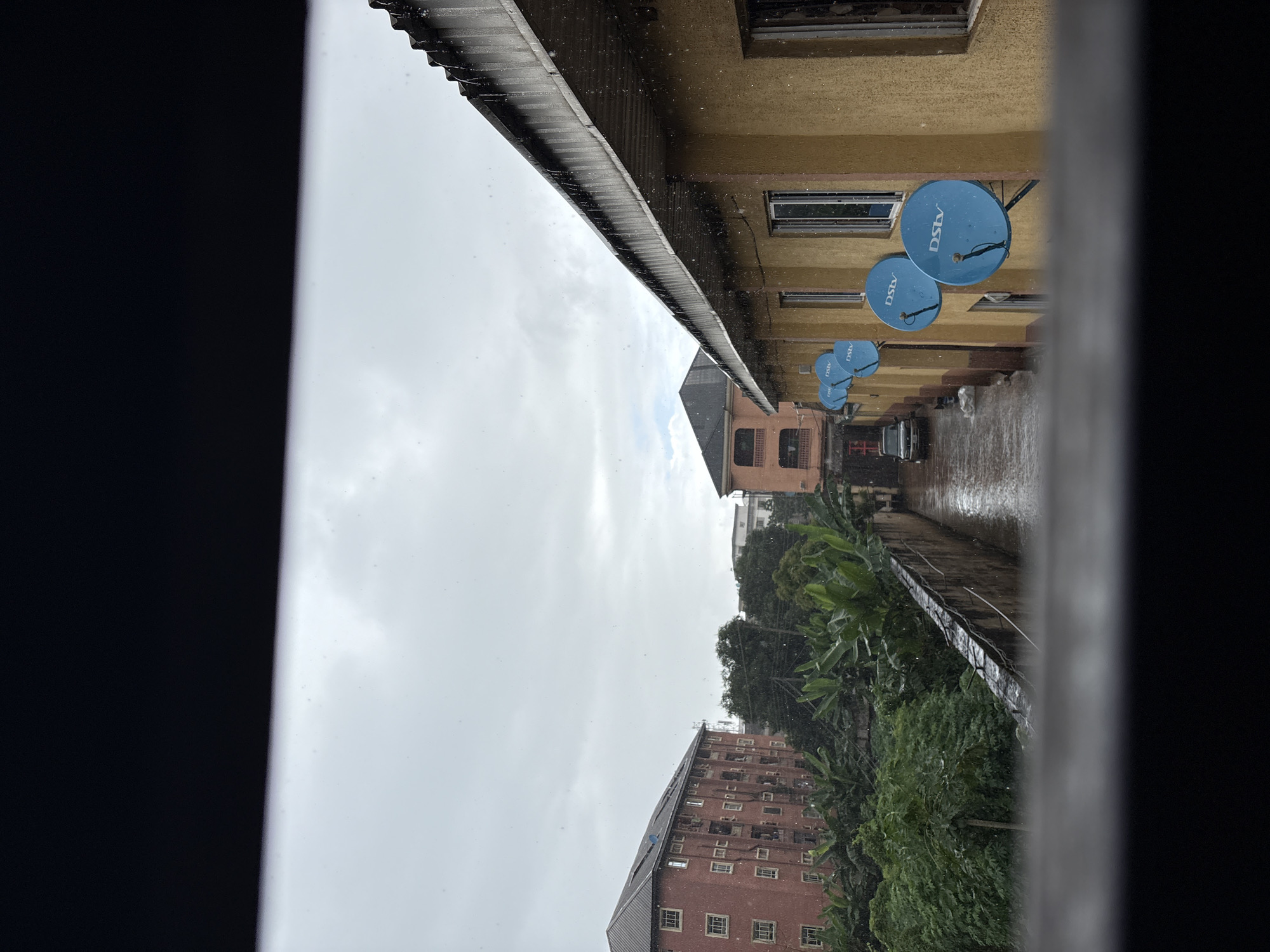 The image depicts a street scene in Onitsha, Nigeria, during a rain shower. The foreground is dominated by a dark, out-of-focus frame, possibly a window or balcony railing, through which the scene is viewed. Beyond this, a residential street is visible with buildings on both sides.
On the right, a two-story building with a yellow-ochre facade occupies a significant portion of the frame. Its corrugated metal roof is visible at the top, with water dripping from the edge, indicating the ongoing rain. Several blue satellite dishes, each with the "DSTV" logo, are mounted on the building's wall. Windows with metal bars are also visible.
To the left of the yellow building, a narrow alleyway or street stretches into the distance. A dark-colored car is parked on the wet, reflective surface of the street, which appears to be flooded in places. The street is lined by a low, concrete wall.
Further to the left, a large, red-brick apartment building stands, its facade covered in windows. Lush green vegetation, including what appear to be banana trees and other foliage, grows in front of and around this building, creating a contrast with the urban architecture. In the background, other buildings and trees are visible under the overcast, rainy sky. The overall atmosphere is one of a typical urban environment experiencing rainfall.