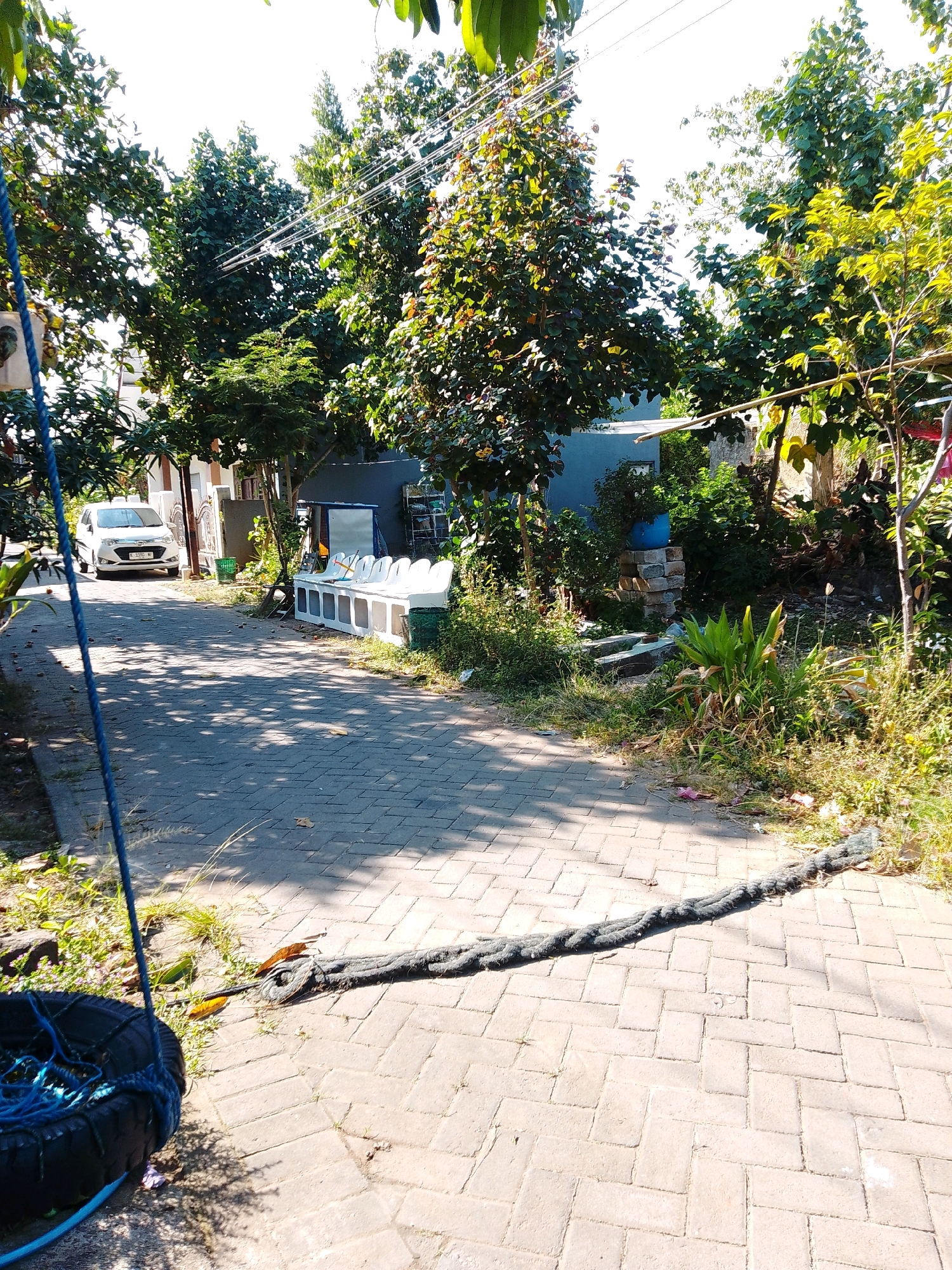 The image depicts a sunlit residential street in Cikal, Indonesia, with a paved walkway leading past houses and lush vegetation. In the foreground, a weathered black tire, suspended by blue ropes from an unseen structure, rests near the walkway's edge. A thick, dark rope stretches across the pavement, anchored to the tire and extending towards the right side of the frame.
To the left of the walkway, a white car is parked in front of a house with a decorative metal gate. Further down the street, a row of white modular seating units is visible, along with some gardening supplies and a blue plastic pot containing a plant. The background features a dense canopy of green trees, partially obscuring the sky and the structures behind them. Power lines are strung overhead, and the sunlight casts strong shadows across the textured pavement, indicating a bright, clear day. The environment appears to be a tranquil neighborhood with a mix of residential housing and abundant plant life.