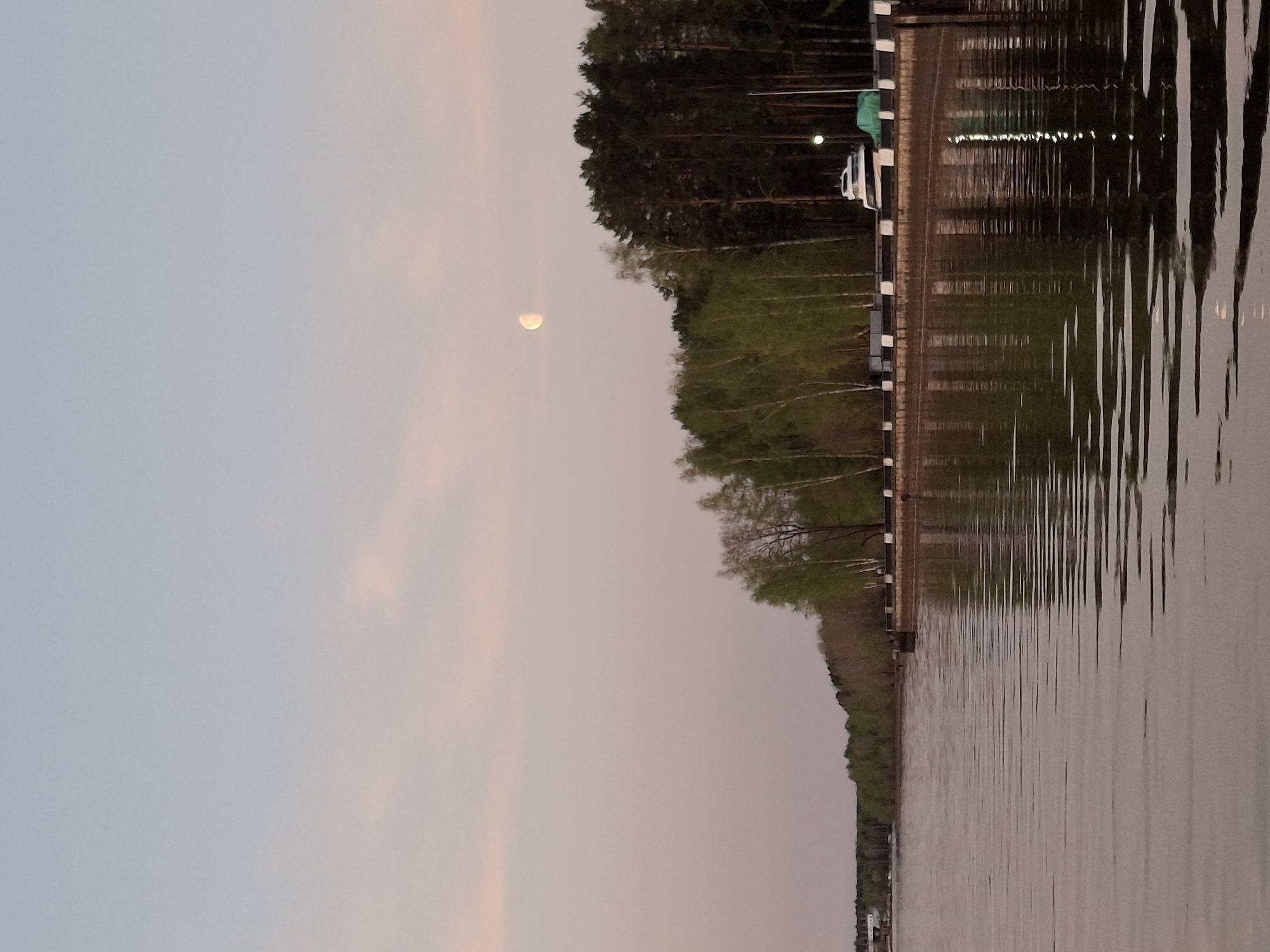 The image captures a tranquil evening scene in Chaikovsky, Russia. A body of water, likely the Kama River due to the city's location, occupies the right half of the frame, its surface reflecting the lights of the shoreline. A line of trees borders the water, transitioning from new, light-green foliage to a darker, dense forest. A walkway with a black-and-white barrier runs along the tree line, where a white vehicle is parked. The left half of the frame showcases a pastel sky, with hues of pink and blue, and a visible crescent moon in the upper left corner. The overall atmosphere is calm and peaceful.