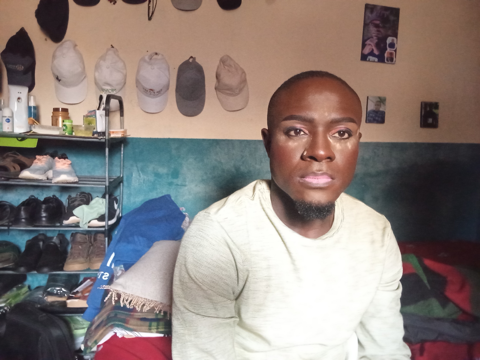 The image depicts a close-up, head-and-shoulders shot of a Black individual with makeup applied, including eyeshadow and lipstick, and a small beard. They are wearing a light-colored, long-sleeved shirt. The background is a wall with several baseball caps hanging on it, and on a shelf, there are various items including what appear to be toiletries and shoes. The scene seems to be set in a room, possibly a bedroom, with a bed visible to the right, covered with blankets. There are also a few framed pictures on the wall. The overall impression is that of a personal space, likely within a residential setting on Rukuba Road, Nigeria.