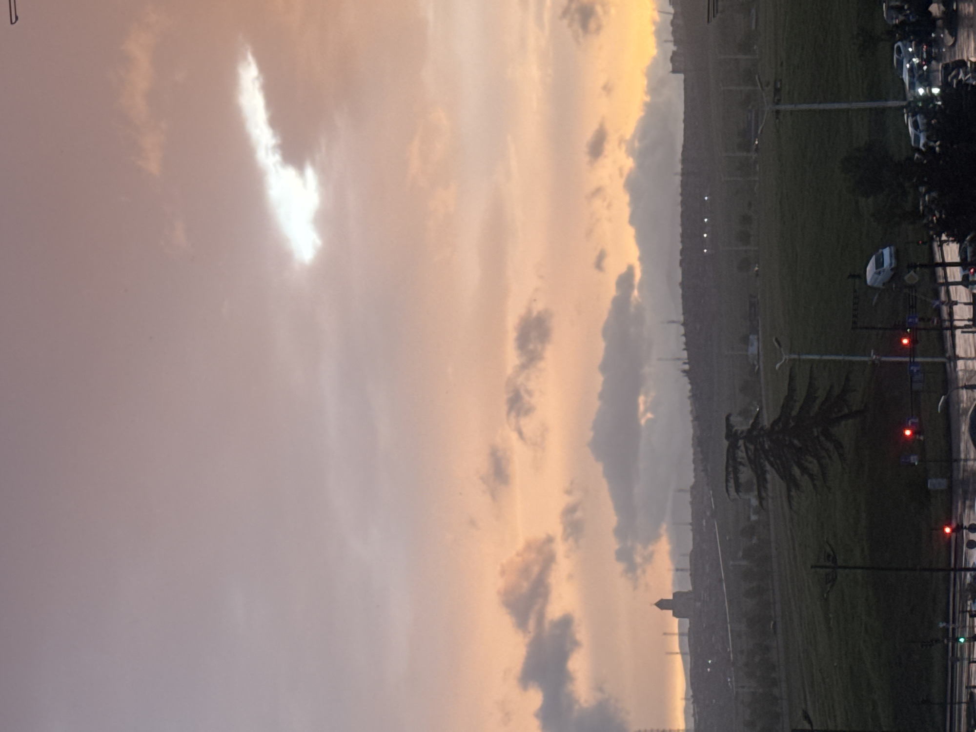 The image shows an early morning scene, likely in Tbilisi, Georgia, based on the visible landmarks and architectural style. The sky is dominated by clouds, illuminated with a peach and grey hue from the rising sun or setting sun. The ground is filled with a green field, and a road with street lights and a few vehicles can be seen to the right. A tall structure, potentially a tower or a monument, is visible in the distance. The foreground includes greenery and what appears to be street lighting fixtures.
