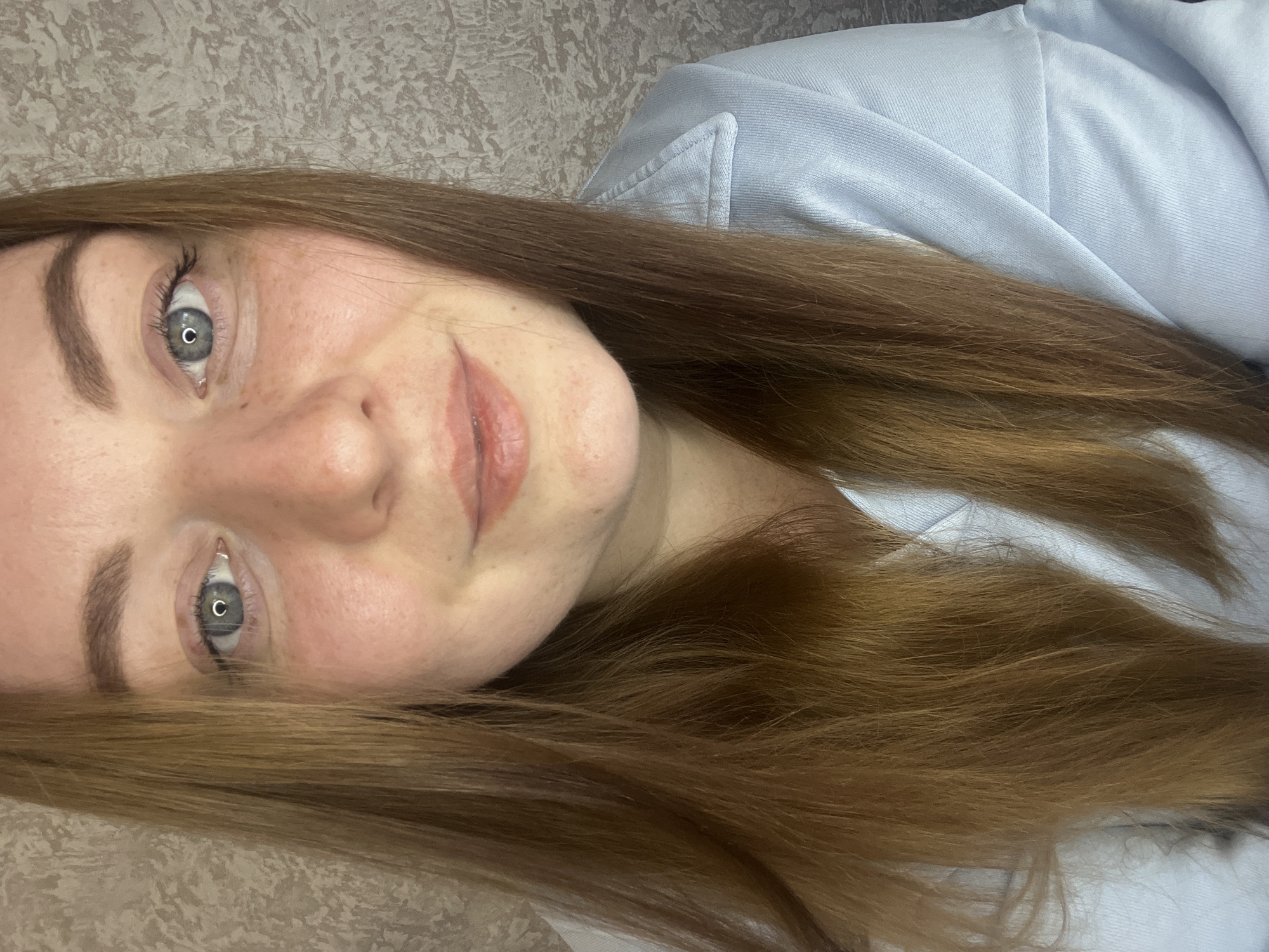 A close-up, portrait-style photograph shows a woman with fair skin, freckles, and light brown hair lying down. Her eyes are a light gray color. She is wearing a light blue top. The background appears to be a textured surface. There is no information to suggest the location of Раздолье, Russia.