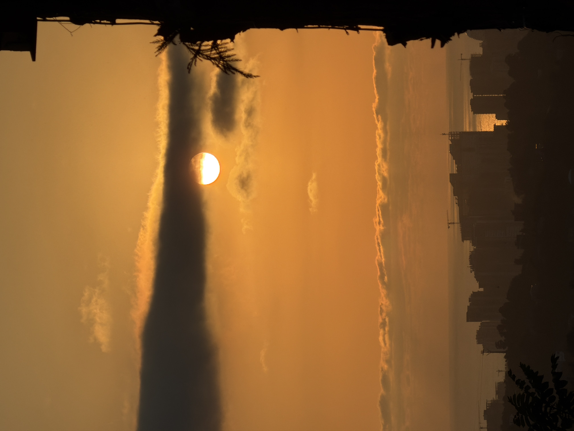 The image captures a silhouetted cityscape against a vibrant orange sunset. The sun, a bright disc partially obscured by dark clouds, is positioned in the upper central part of the frame, casting a warm glow across the sky. Below, a layer of thinner clouds is illuminated by the setting sun. In the foreground, on the right side, a tall, dark, silhouetted structure, possibly a building or a tree, dominates the edge of the frame. Further in the background, a city skyline is visible in silhouette, with various buildings and construction cranes dotting the horizon. The sea is faintly discernible beyond the city. The overall impression is one of a serene, twilight urban landscape.