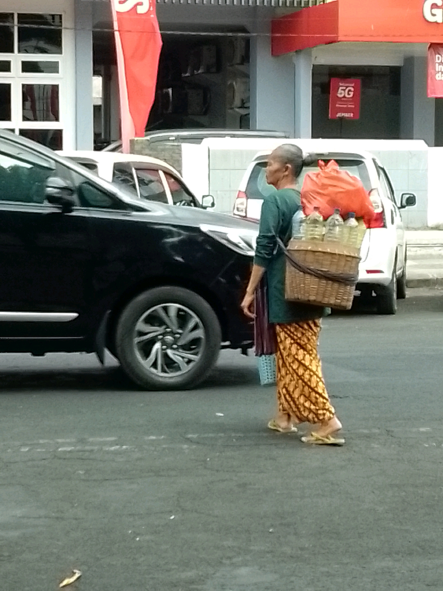A person is walking on a street in Jember, Indonesia, carrying a basket of bottled water on their shoulder. The person is wearing a green long-sleeved shirt, a patterned skirt, and sandals. The basket contains multiple plastic bottles, and a large orange bag is tied to the top of the basket. A black car is parked on the left side of the frame, and a white car is visible behind the person. In the background, there is a building with a red awning and signs, one of which displays "5G". The street appears to be paved, and there are some scattered debris on the ground.