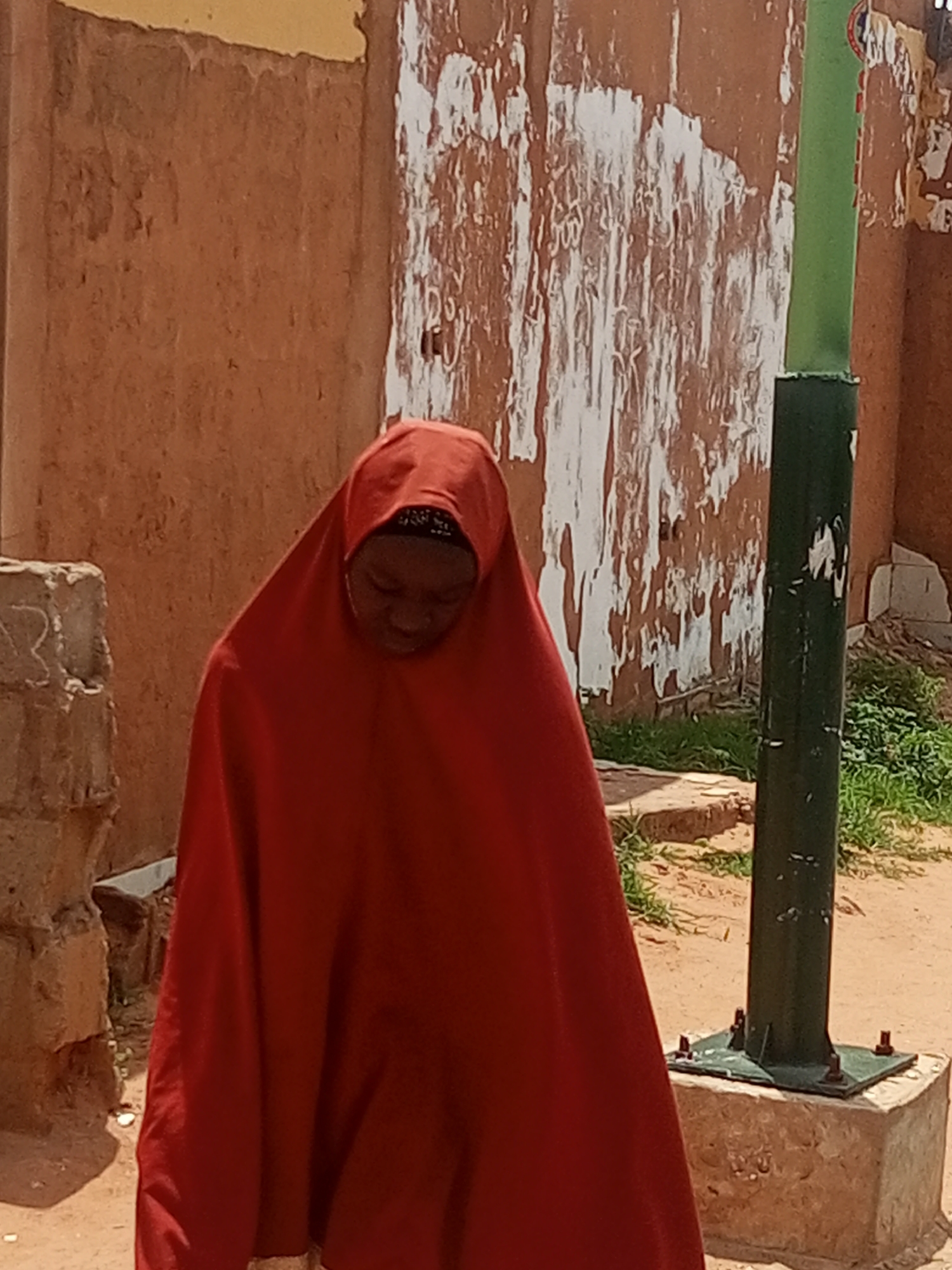 A person wearing a bright red hijab is shown from the chest up, with their head bowed. They appear to be looking down, possibly in thought or prayer. The backdrop consists of a textured, light brown wall with patches of peeling white paint, suggesting an aged or weathered structure. To the right of the person is a dark green pole, possibly a lamppost, mounted on a concrete base. Green foliage and dry ground are visible around the base of the pole and in the background. The lighting suggests it is daytime, with sunlight casting shadows. The scene appears to be outdoors in a possibly arid or dusty environment, given the sand-colored ground. No text is visible in the image. The emotion conveyed is one of quiet introspection or humility.