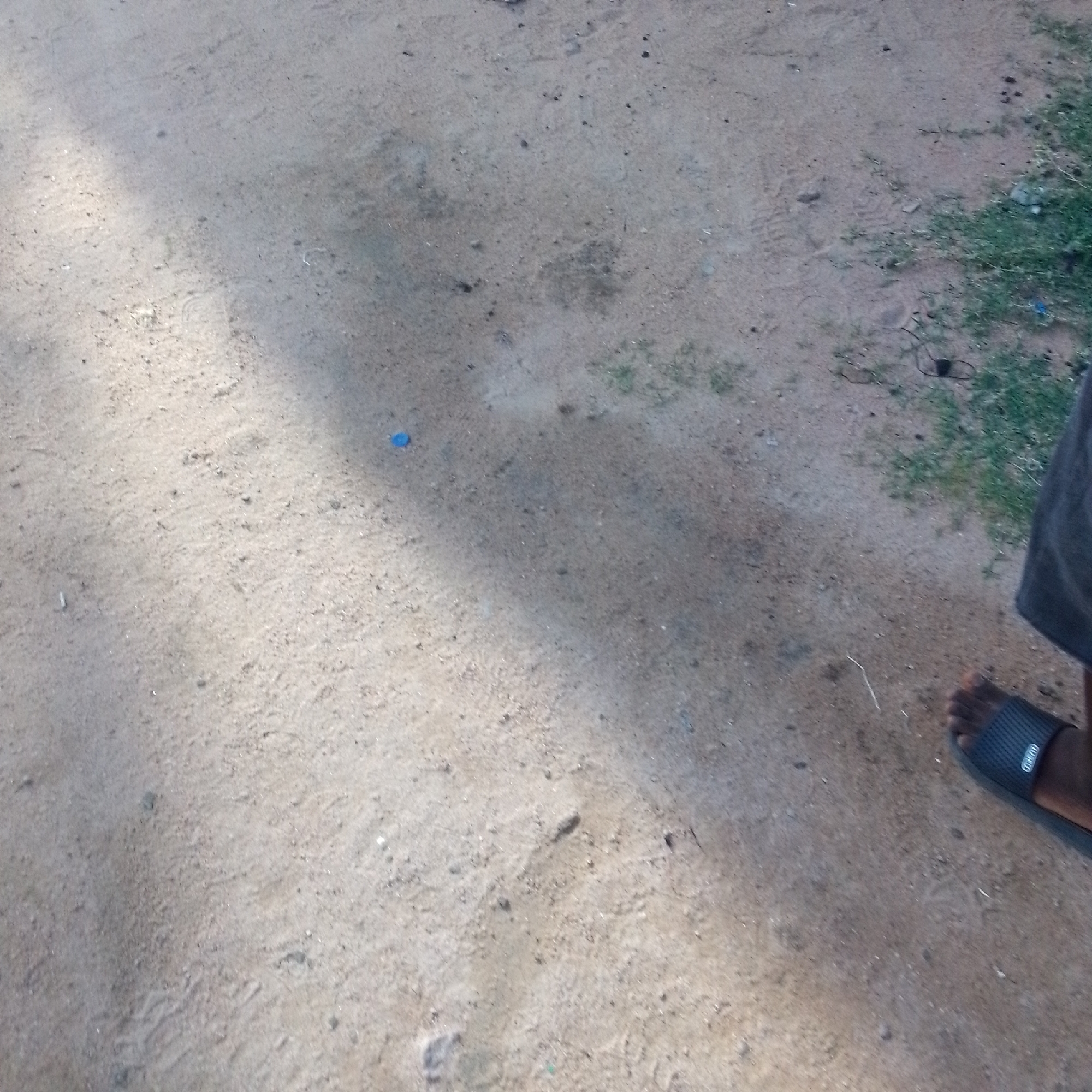 An overhead, medium shot shows a person's foot wearing a black sandal against a sandy, outdoor ground. The ground appears to be a mixture of dirt and small pebbles, with some patches of dry grass. The lighting suggests it is daytime, with a shadow cast across the ground from the left side of the frame. A small, blue object, possibly a bottle cap, is visible on the ground to the left of the center. The person's foot is positioned in the lower right corner of the frame, with only the sandal and toes clearly visible. The rest of the person's body is out of frame.