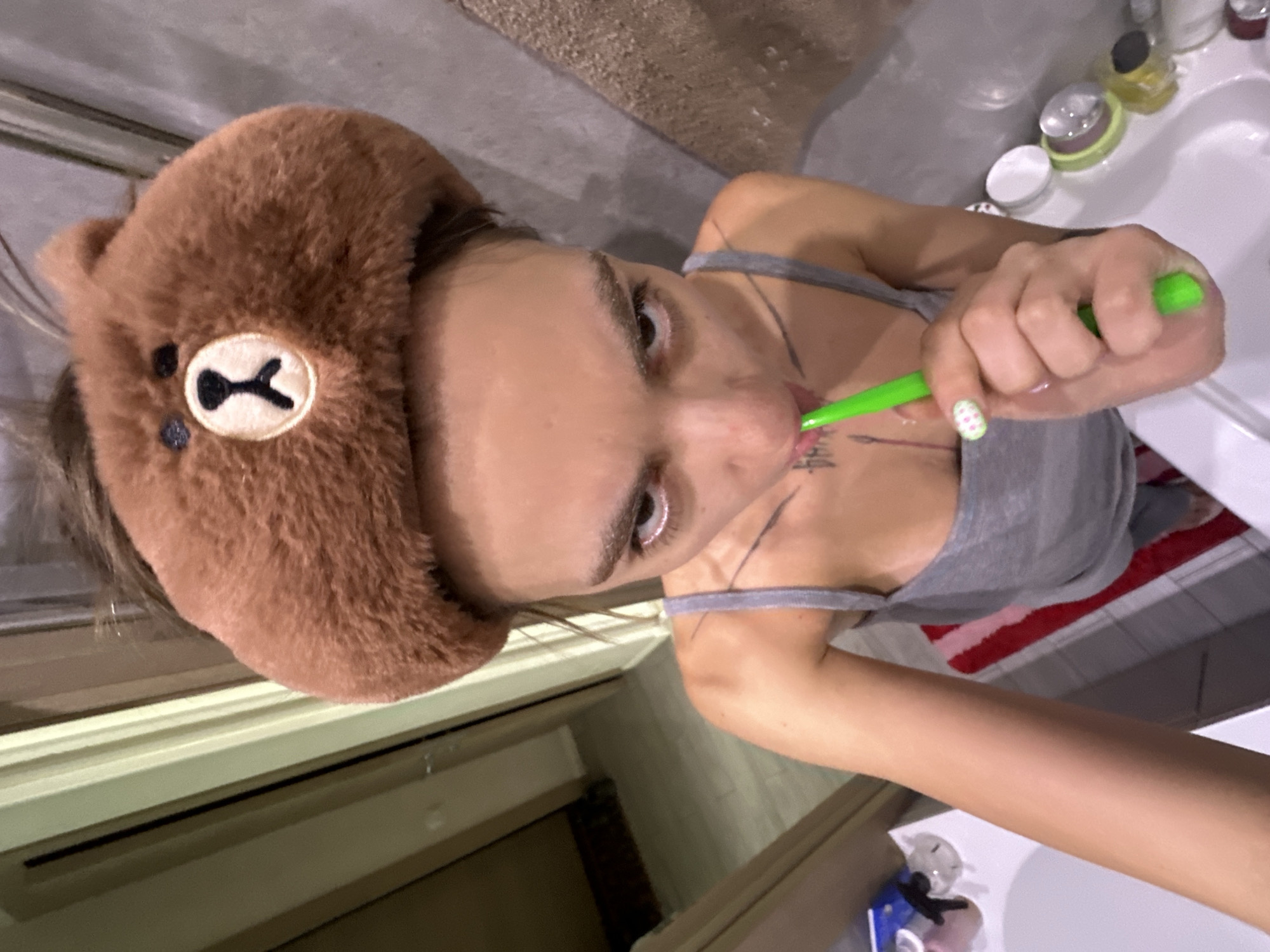 A person is brushing their teeth in a bathroom. They are wearing a brown fuzzy headband shaped like a bear's head. The person is holding a green toothbrush and appears to be looking into a mirror. The bathroom appears to be somewhat cluttered, with various toiletries visible on the counter. The overall lighting is dim. This scene is not identifiable as being in Moscow, Russia.