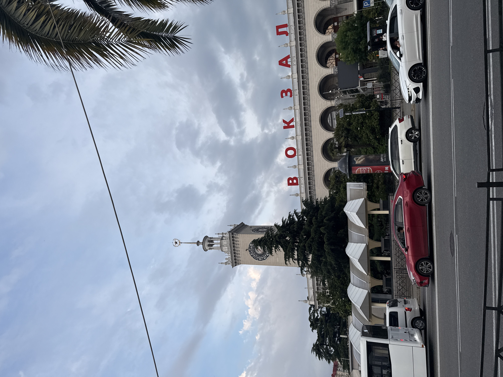 This is a medium shot of the Sochi train station in Sochi, Russia, on a cloudy day. The large, imposing building features a prominent clock tower with a star atop it. Red letters spelling out "ВОКЗАЛ" (which translates to "Station") are visible across the top of the building. Several vehicles, including a white bus, a white van, a red sedan, and a white sedan, are driving on a multi-lane road in the foreground. Lush green trees are planted in front of the station. The sky is filled with dramatic, grey clouds, and a palm frond is visible in the upper right corner of the frame.