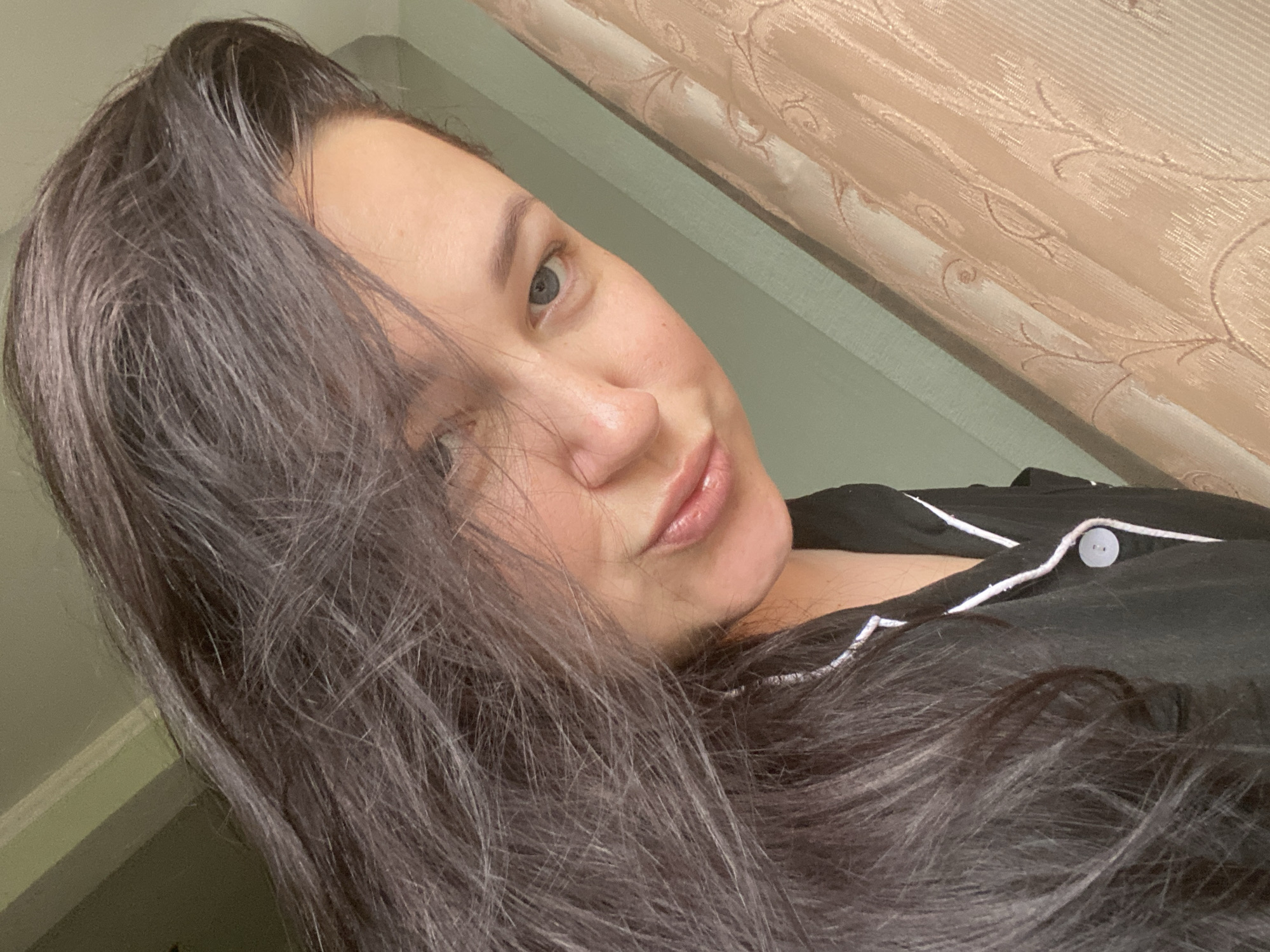 A woman with long, dark hair is looking at the camera with a slight smile. She has light skin and blue eyes. She is wearing a black pajama top with white piping and a button closure. The background includes a light-colored curtain and a light green wall. There is no visible indication that the image was taken in Sochi, Russia.