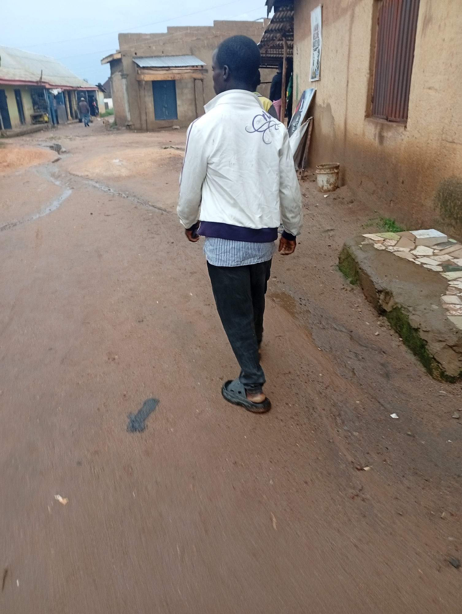 In Dong, Nigeria, a solitary individual walks away from the camera down a wet, reddish-brown dirt road. The person is wearing a white jacket with purple decorative stitching on the back, a blue and white striped shirt, black pants, and grey Croc-style sandals. The road is lined with simple, weathered buildings made of concrete blocks and corrugated metal roofing. Some buildings have blue doors and windows. In the background, other individuals can be seen walking or standing near the structures. The sky is overcast, contributing to a muted lighting condition. The environment appears to be a residential or commercial area in a developing region, characterized by informal construction and unpaved roads.
