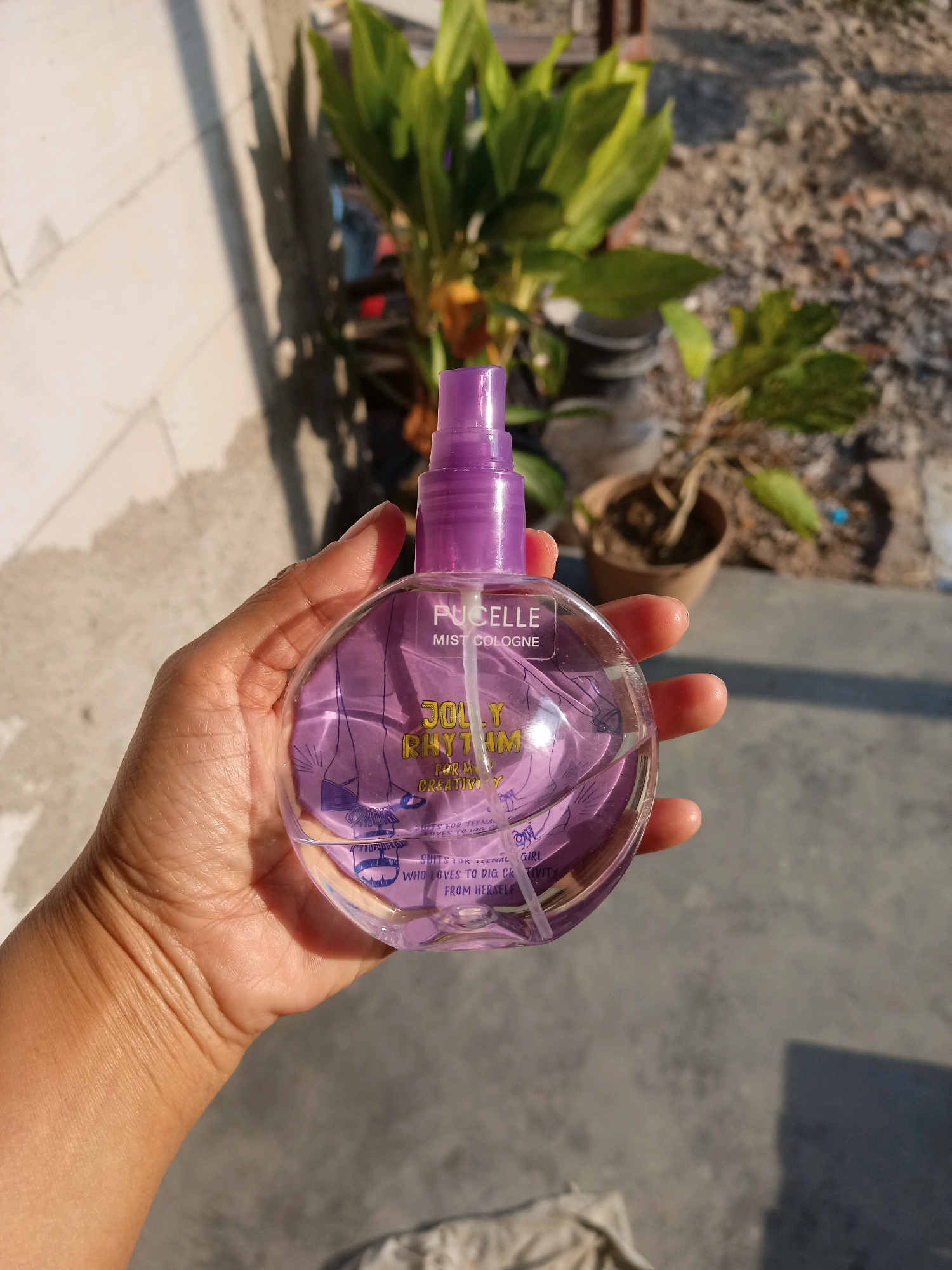 A person's hand holds a round, clear bottle of Pucelle Mist Cologne, brand "JOLLY RHYTHM" in purple lettering, with a purple spray nozzle. The bottle contains a purple liquid and has illustrations on the label. The background is outdoors, with a white brick wall casting a shadow on the left, and gravel and potted plants in the background. The scene appears to be in Karangtalun, Indonesia, given the context. The lighting suggests it is daytime.