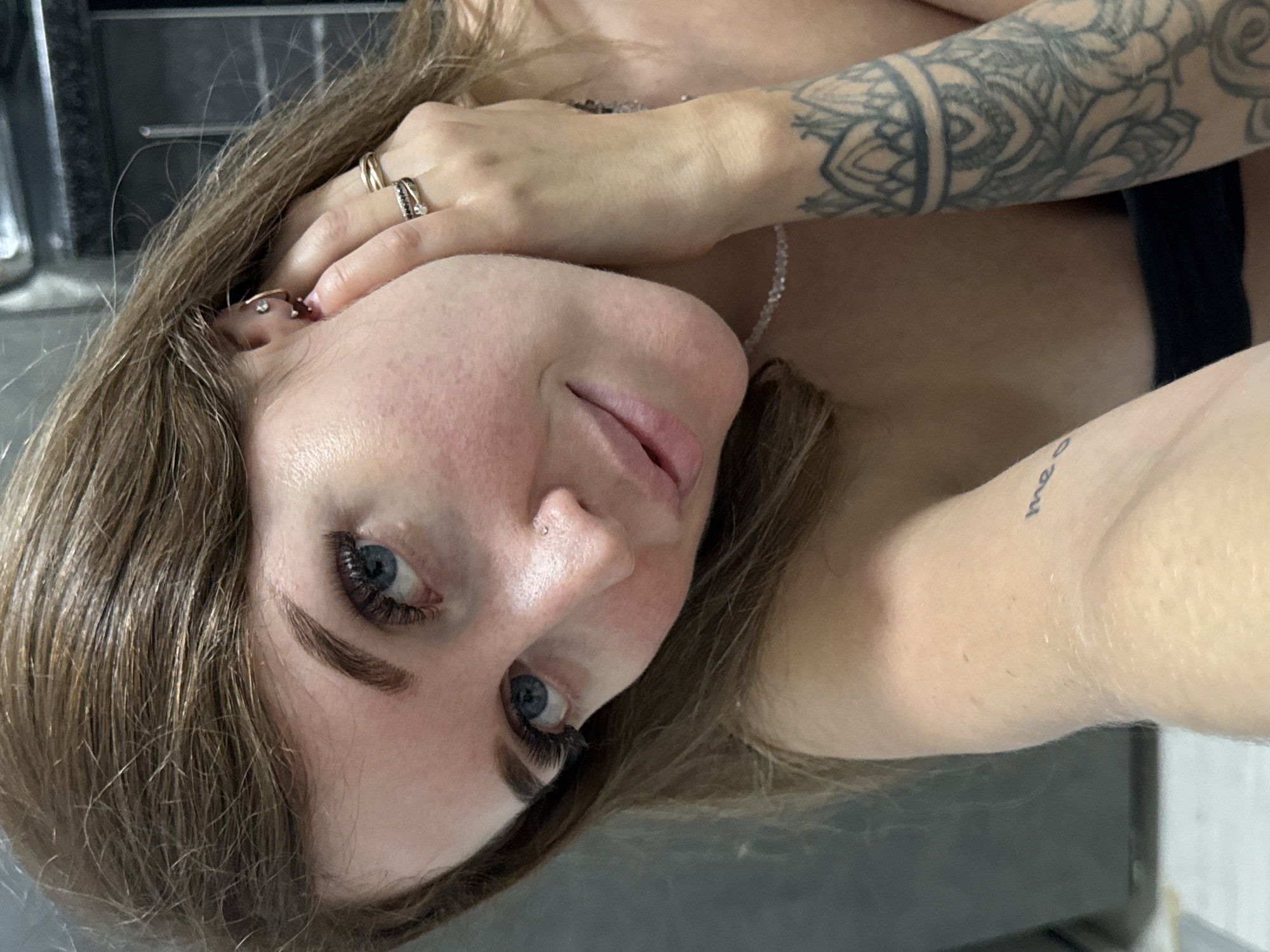 A medium shot photograph shows a young woman with brown hair and blue eyes posing for a selfie. She is wearing a black strapless top and has several pieces of jewelry, including rings on her fingers, earrings, a nose ring, and a necklace. She also has tattoos on her arm. The background appears to be indoors with a dark gray wall. The image does not contain any information that would indicate its location in Chaikovsky, Russia.