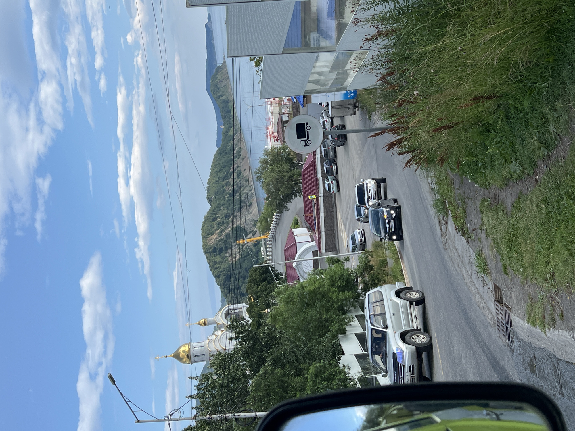 The image captures a scene in Petropavlovsk-Kamchatsky, Russia, likely from within a vehicle. In the foreground, the side mirror of a light-colored vehicle is visible on the left, reflecting a green hue. A silver SUV is in motion on the road, followed by a black sedan. The road curves to the right, and on the right side of the road, a traffic sign depicting a truck is prominently displayed. Beyond the road, a large, white building with golden domes, identified as the Church of the Resurrection, stands out against a backdrop of lush green hills and a blue sky with scattered clouds. In the distance, a body of water, likely Avacha Bay, is visible with a port or industrial area along its edge. The environment appears to be a coastal city with a mix of urban infrastructure and natural landscapes.