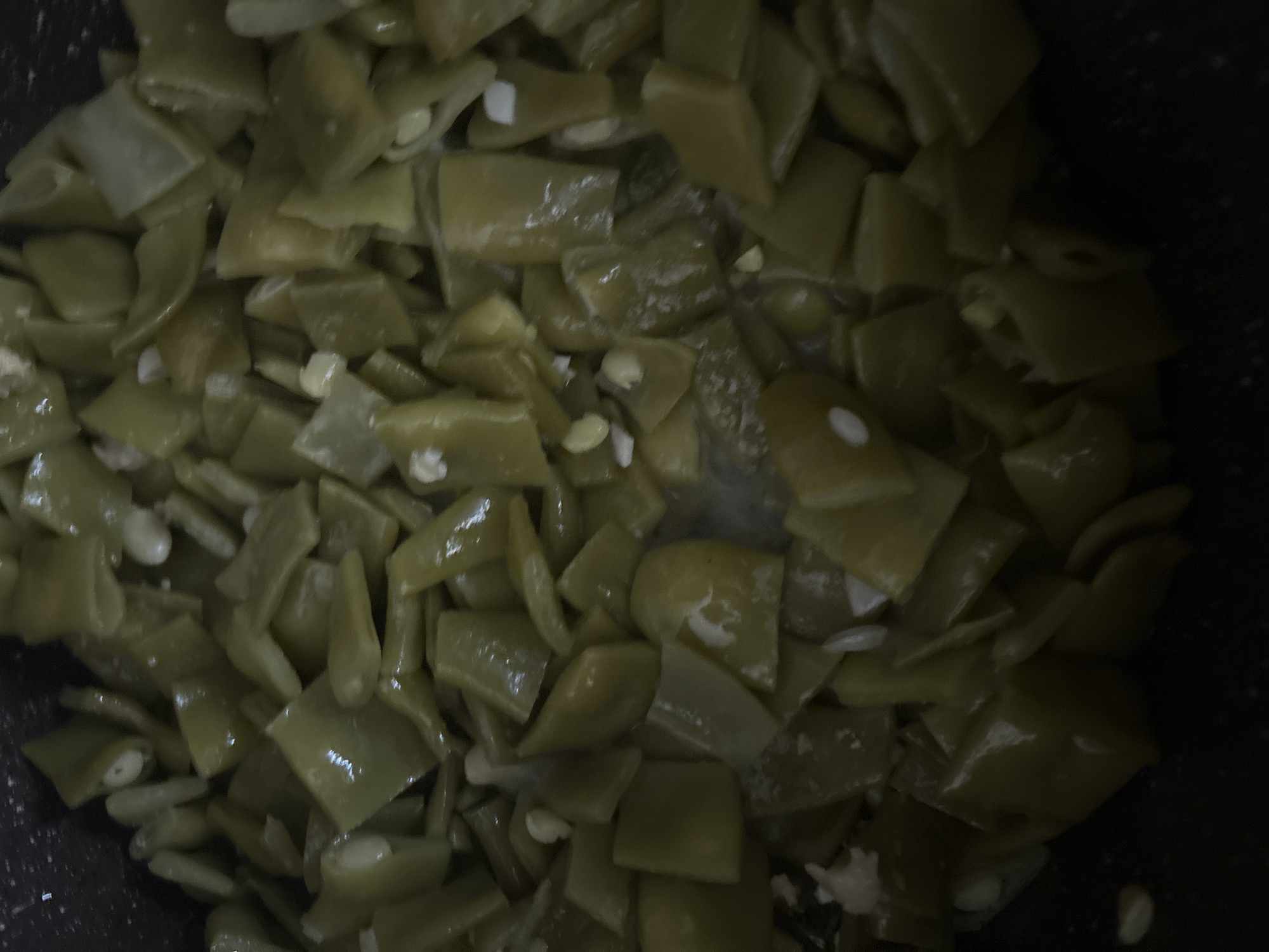 A close-up, overhead shot shows cooked green beans in a dark pot. The beans are chopped into bite-sized pieces and appear to be glistening, indicating they may be in a sauce or have been cooked with oil. Some white specks, possibly garlic or onion, are visible amongst the beans. The lighting is dim, casting shadows and making it difficult to discern further details of the environment. The image is cropped to focus solely on the food.