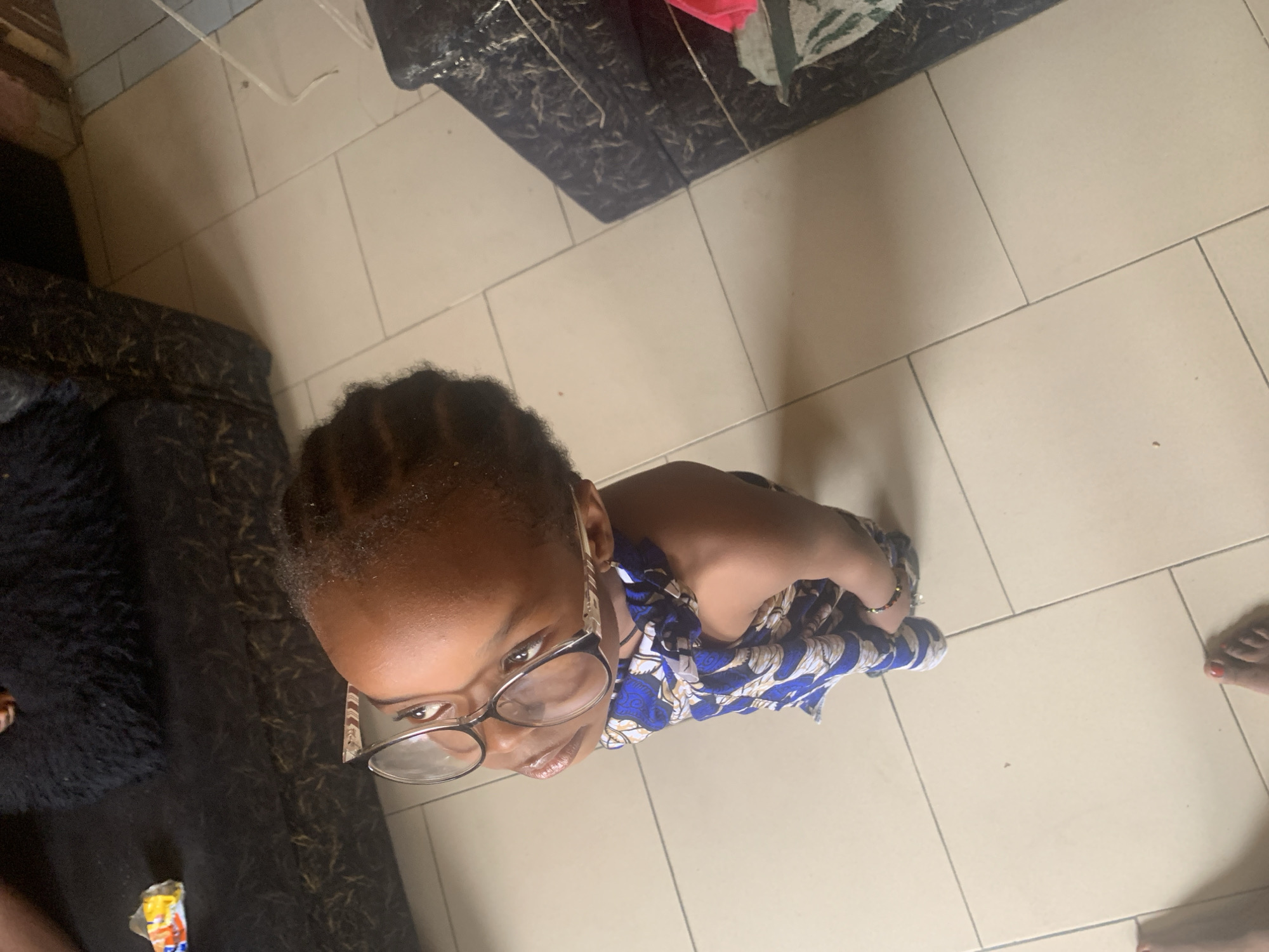 A high-angle indoor shot shows a young dark-skinned child wearing glasses and a blue and white patterned dress. The child is standing on a tiled floor and looking up at the camera. To the left of the child is a black furry object, possibly a rug or piece of furniture, and a partially unwrapped candy or snack. In the top left corner, there is a white cord. In the background are pieces of furniture with patterned upholstery. A glimpse of an adult's foot is visible in the bottom right corner. The location is not indicated in the picture, however, it is indicated that the photograph was taken in the city of Sabon Tasha, Nigeria.