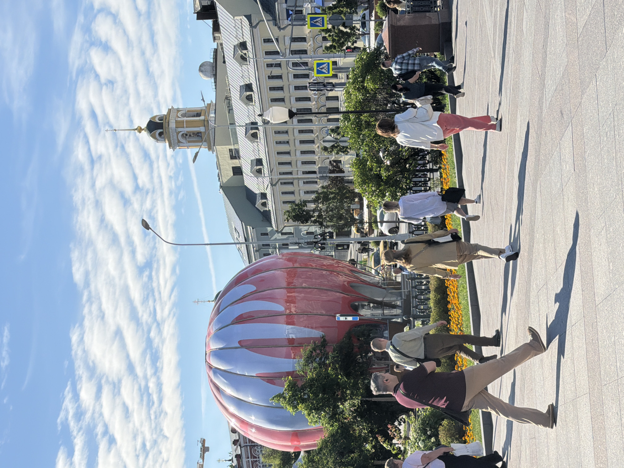 The image shows a street scene in Moscow, Russia, on a sunny day with a clear blue sky and scattered white clouds. Several people are walking on a paved walkway lined with flowerbeds and trees. In the background, there is a large, modern red and silver spherical structure, possibly an art installation or exhibit. Behind it, a traditional building with a domed tower and intricate architectural details is visible, suggesting a mix of historical and contemporary elements in the city.
One person in the foreground is wearing a dark red shirt and light brown pants, walking with a purposeful stride and casting a long shadow. Another person further back is wearing a white top and pink pants, also walking on the pavement. Several other individuals are walking in the same direction. The overall atmosphere appears to be casual and lively, with people enjoying a day outdoors. The lighting suggests it is daytime, likely late morning or early afternoon, given the strong shadows. There is no visible text in the image.