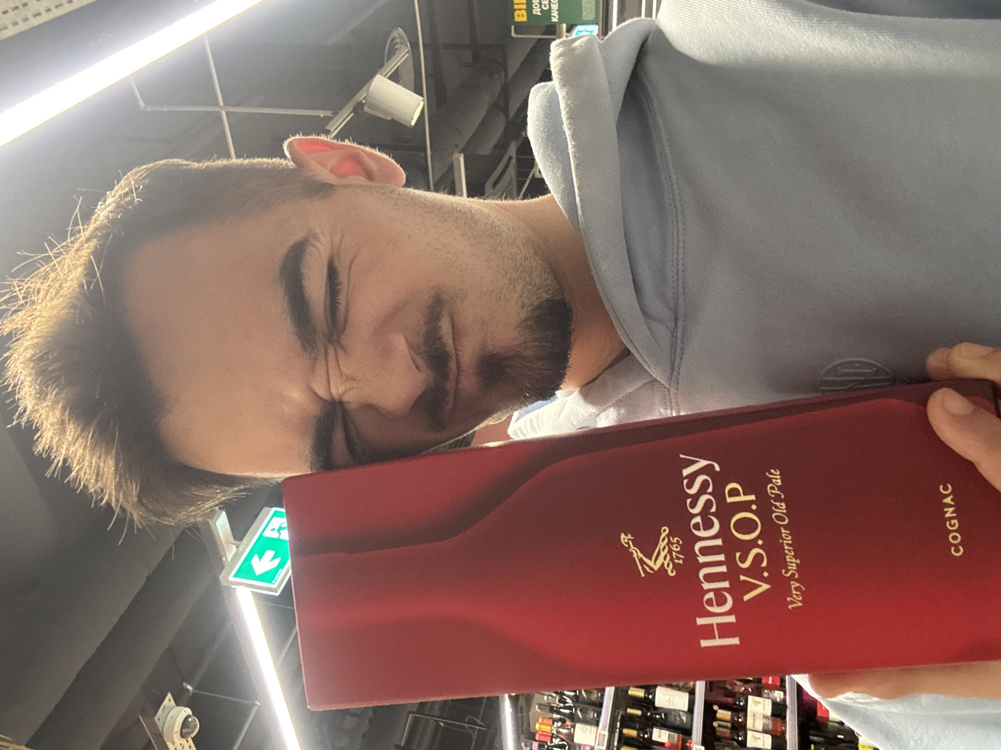 A man, likely in his 20s or 30s, is depicted holding a red box of Hennessy V.S.O.P. Very Superior Old Pale Cognac. He wears a light blue hooded sweatshirt. His face is turned towards the camera, with eyes tightly squinted and a somewhat grimacing or playful expression, possibly reacting to the bright overhead lighting. He has short brown hair and a light beard. His right hand is visible, gripping the Hennessy box.
The scene is indoors, strongly suggesting a supermarket or liquor store aisle in Sofia, Bulgaria. This is indicated by the background elements: shelves stocked with various bottles, bright fluorescent lights on the ceiling, exposed pipes, and a security camera. A green sign with a white downward-pointing arrow, commonly an exit indicator, is visible behind his head. Further in the background, a sign with Cyrillic text can be seen, which appears to include "БИЛА" (BILLA), a supermarket chain prevalent in Bulgaria.
The Hennessy box prominently features its brand name, "V.S.O.P. Very Superior Old Pale," "COGNAC," and the establishment year "1765" in gold lettering. The overall atmosphere is well-lit, typical of a commercial retail environment. The time of day is not discernible due to the indoor setting and artificial lighting.