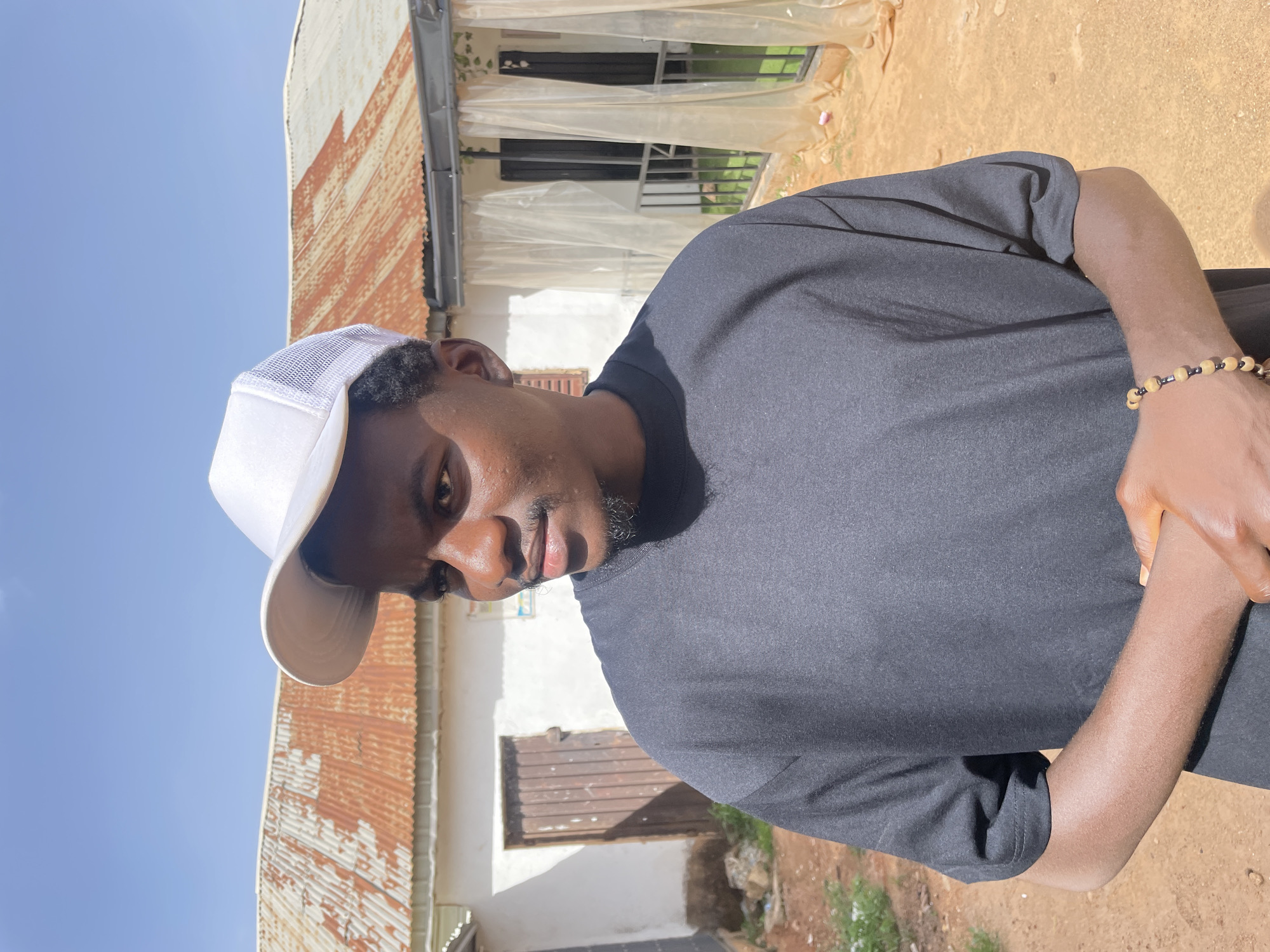 The image features a dark-skinned male wearing a white baseball cap and a dark t-shirt. He is standing outdoors, with a building in the background composed of white walls, brown doors, and rusty corrugated metal roofing. A window is visible behind a white plastic sheet. The ground is reddish-brown soil. The sky is blue. A beaded bracelet is visible on his left wrist. The location is likely Barakin Akawo, Nigeria, based on the prompt. The subject is looking downwards.