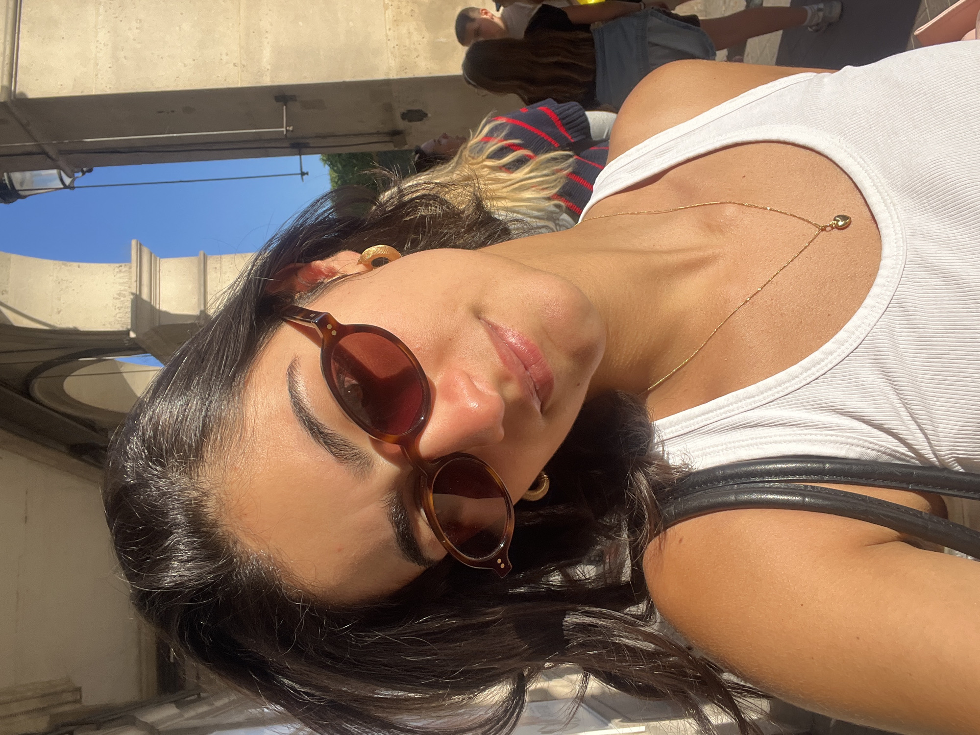 The image is a close-up selfie of a young woman taken outdoors on a sunny day. She has dark hair and a tanned complexion, wearing a white ribbed tank top and stylish dark sunglasses with tortoiseshell frames. A delicate gold chain necklace with a small heart pendant adorns her neck. A subtle smile is visible on her lips beneath the sunglasses. A black bag strap is partially visible over her shoulder.
The selfie is framed from a low angle, looking upwards towards a bright blue sky that is visible between concrete architectural structures, possibly parts of bridges or elevated walkways. In the blurred background, other individuals are partially visible, including one person with long blonde hair wearing a striped top (red and dark blue/black).
The strong, natural sunlight indicates it is daytime, likely midday or afternoon, contributing to a bright and casual atmosphere. The presence of architectural structures and other people suggests a public urban environment, consistent with the provided location context of Paris, France. No text is visible in the image.
