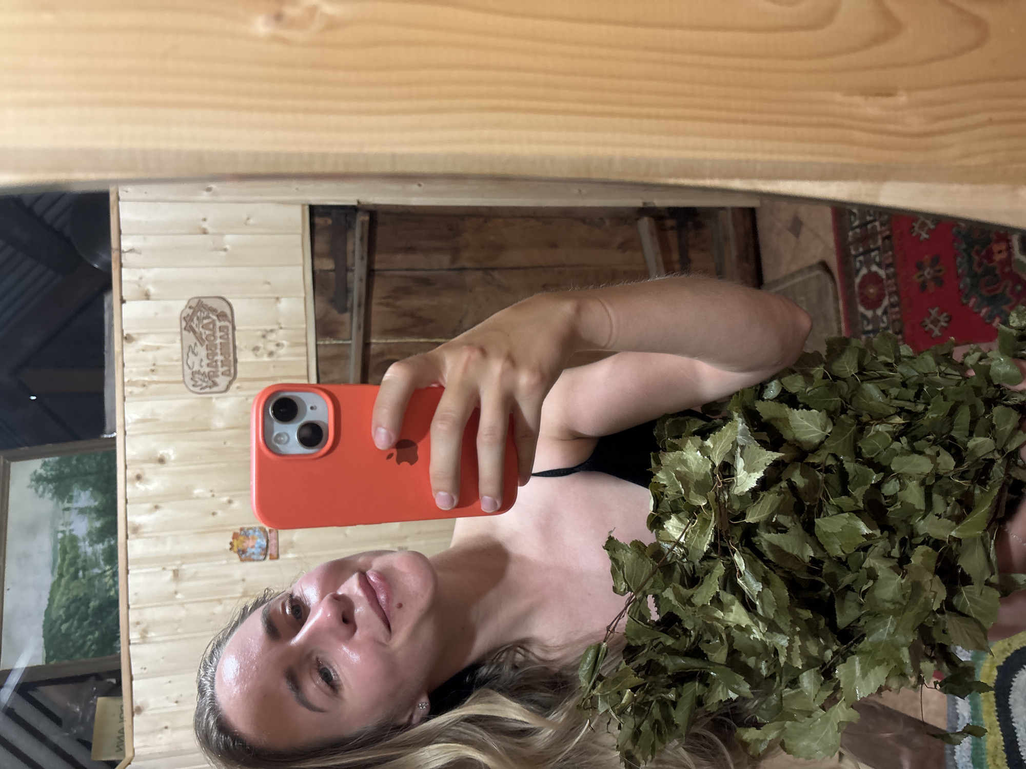 A fair-skinned woman with blonde hair pulled back slightly is taking a selfie in a wooden sauna. She is wearing a black tank top and holding a bunch of green leaves, possibly a venik, in her left arm. Her right hand is holding an orange smartphone up to take the picture. The background shows light-colored wooden walls, a darker wooden door on the right, and a painting of a landscape on the upper left wall. The overall lighting suggests the interior of a dimly lit room. The location is identified as Ваньки, Russia.