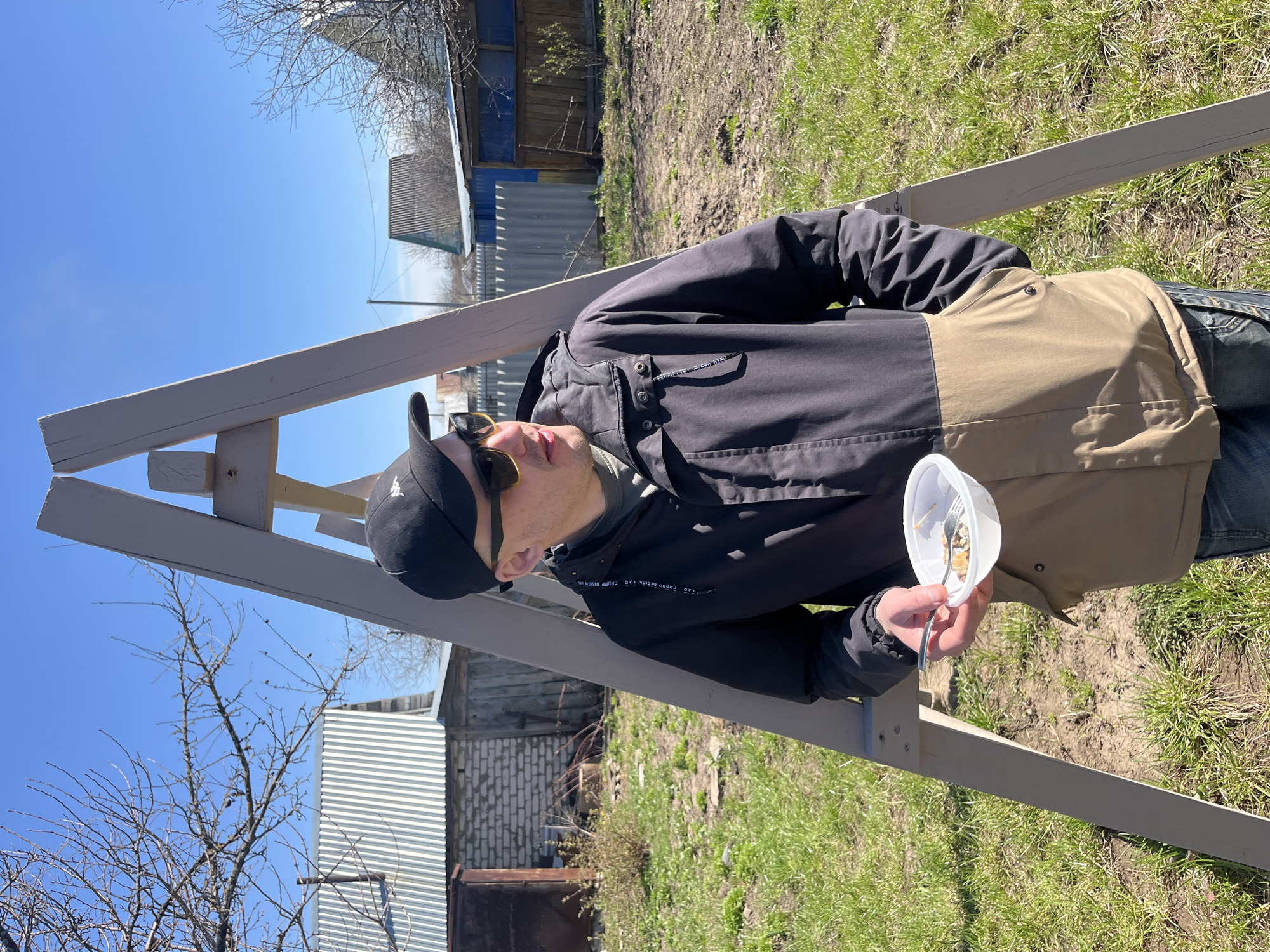 A man stands outdoors in Neftekamsk, Russia, holding a white container with food and a fork. He wears a black baseball cap, sunglasses, and a black jacket with a beige section. He is leaning against a light-gray wooden structure. The background includes green grass, a fence, a tree without leaves, and residential buildings. The sky is blue.