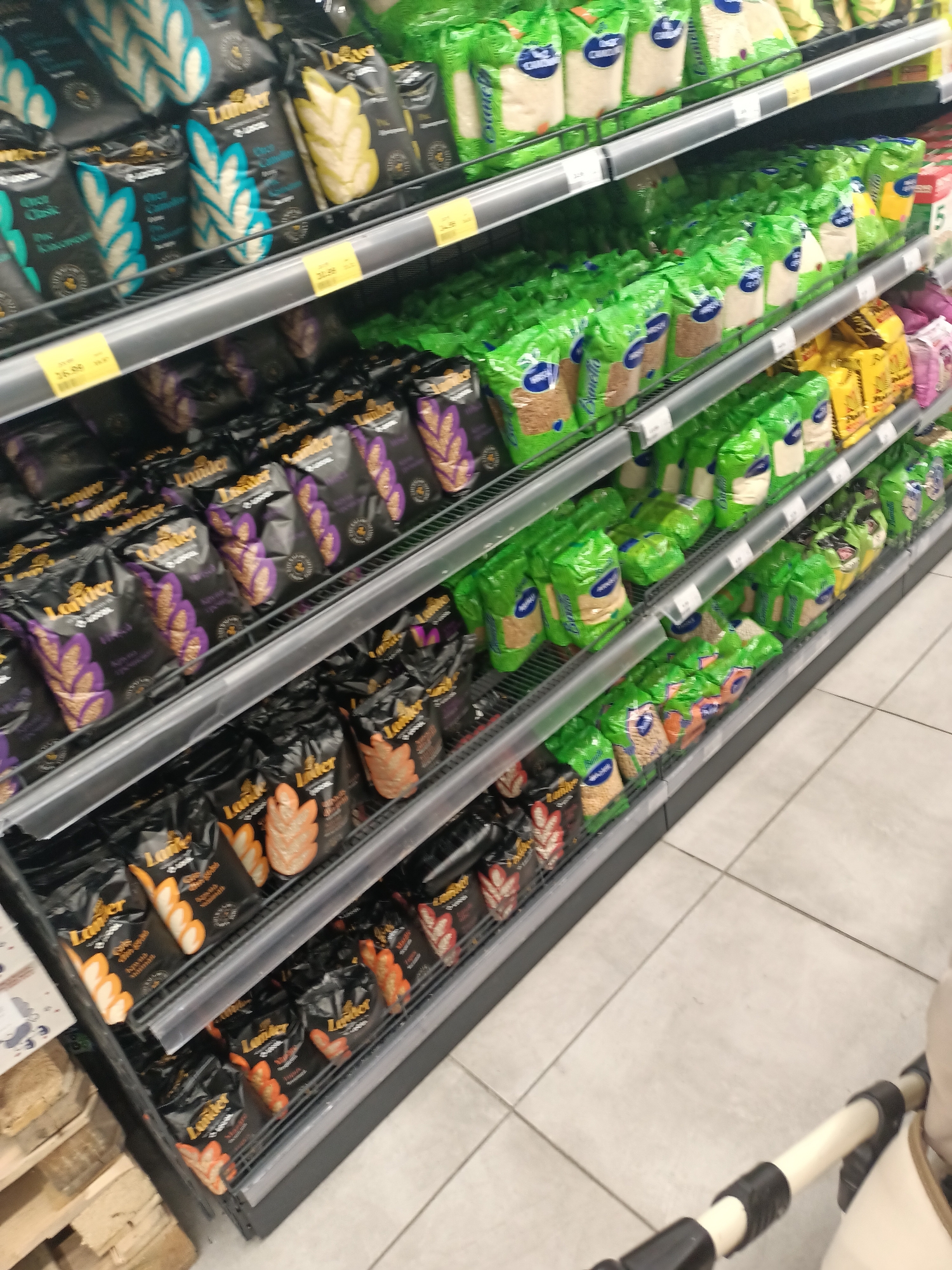 The image shows a retail store aisle with shelves stocked with packaged food items, primarily bags of grains and cereals. The shelves are arranged vertically, and the products are neatly displayed. The lighting is bright and even, typical of a supermarket. On the right side of the image, a portion of a baby stroller is visible, suggesting the presence of shoppers. The floor is tiled in a light gray color. The overall impression is a well-organized and brightly lit retail environment.
