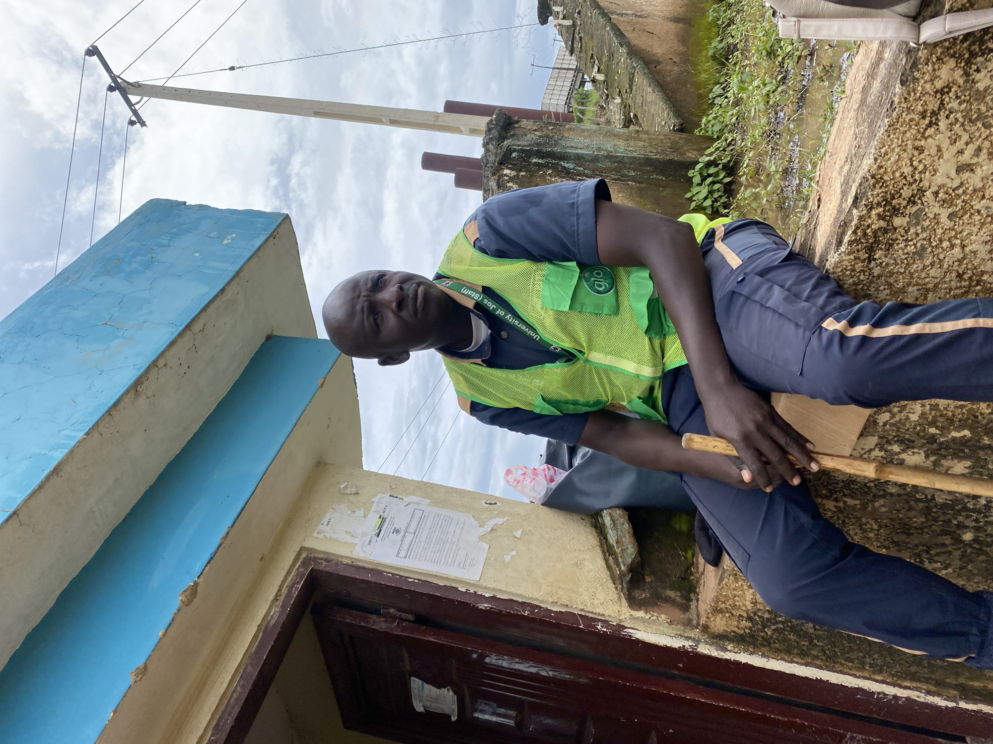 In Rafin Par, Nigeria, a bald Black man wearing a dark blue polo shirt with a bright green high-visibility vest over it is seated on a concrete ledge. The vest has "University of Jos (Staff)" printed on a band around his neck and a "Glo" logo on the chest pocket. He holds a light-brown wooden stick in his hands. The man is positioned in front of a building with a light yellow facade and a blue trim under the roof. A dark-colored door is visible to his left. Papers are tacked to the wall next to the door. The ground in front of the ledge is muddy and overgrown with green vegetation, suggesting it might be a rainy season or an area prone to flooding. In the background, a utility pole with wires is visible against a cloudy sky.
