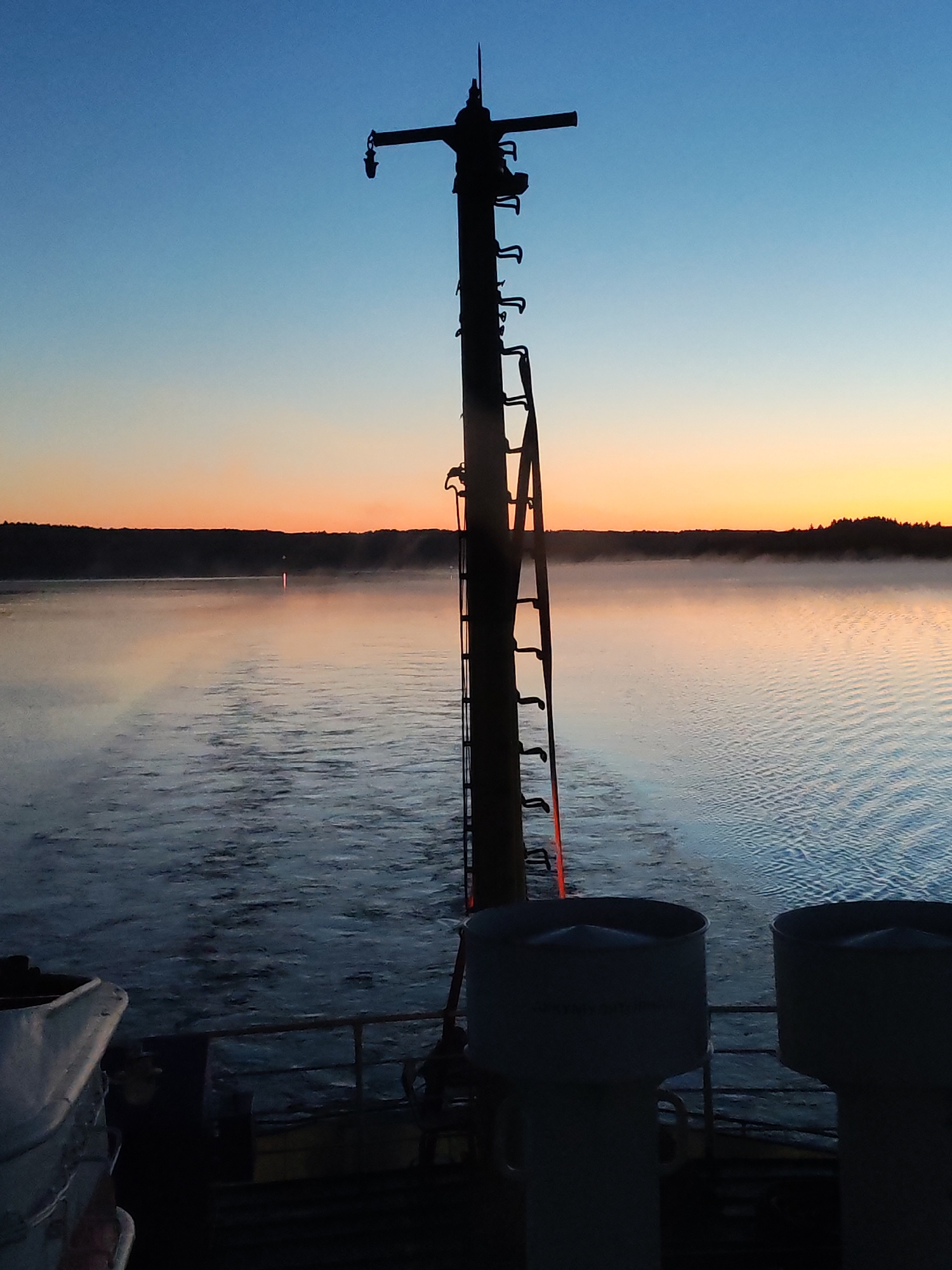 This image captures a pre-dawn or twilight scene from the stern of a vessel on a large body of water, located in the region of Irma, Russia. The sky exhibits a clear gradient, transitioning from a deep twilight blue at the top to a lighter blue mid-sky, and then to vibrant orange and yellow hues closer to the horizon.
A dark, silhouetted landmass, consistent with a forested shoreline, extends horizontally across the distant horizon. The surface of the water in the mid-ground is covered with a layer of atmospheric mist or fog, which appears to reflect the orange light from the sky. A distinct vertical streak of orange-pink light is visible on the water's surface in the distance, likely a reflection of the rising or setting sun or a distant light source. The water immediately behind the vessel shows a clear wake and ripples, indicating the boat is in motion.
In the foreground, a tall, dark, mast-like structure rises centrally from the vessel's deck, casting a prominent silhouette against the sky. This mast is equipped with a vertical series of rungs or hooks resembling a ladder, and near its top, a horizontal arm extends to the left, suspending a dark, downward-facing lantern or light fixture. A faint reddish-orange glow is present on the lower section of the mast and on a dark railing situated below it.
To the bottom left, a section of a white, curved object, possibly a life raft container or similar deck equipment, is visible. To the bottom right, two large, dark, cylindrical structures, appearing to be ventilation shafts or exhaust funnels, rise from the deck. No individuals are visible in the frame.