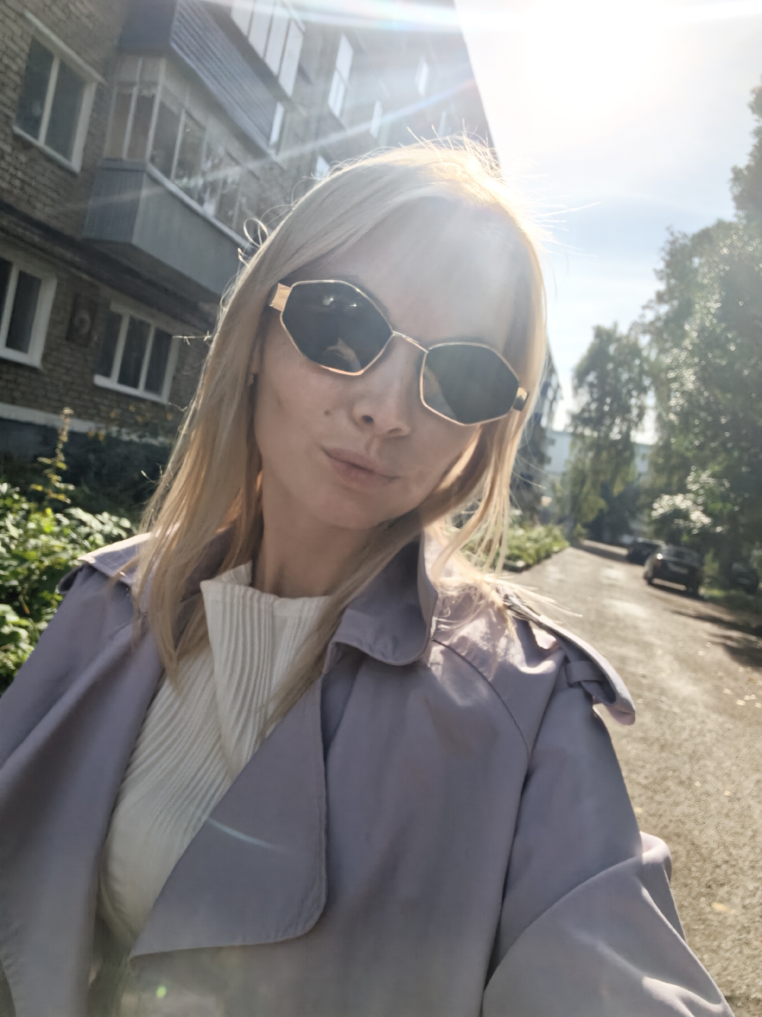 A blonde woman with sunglasses is taking a selfie outdoors. She is wearing a light-colored pleated top and a lavender trench coat. The background shows a brick building and trees, with a road and parked cars in the distance. The lighting suggests it is daytime with sunlight. The image appears to be taken on a street in the city of Sterlitamak, Russia.