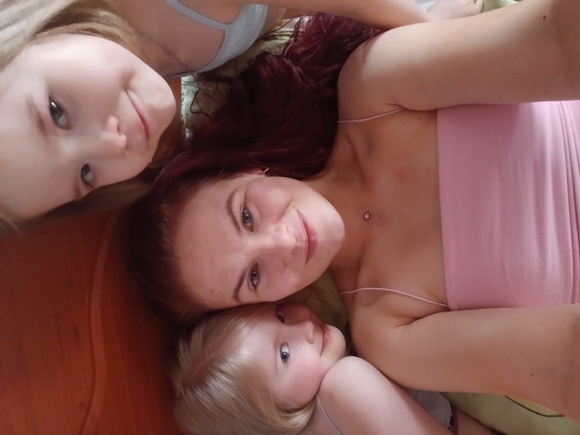 The image shows a selfie taken by a woman with two young girls. The woman has auburn hair and is wearing a pink top. The girls both have blonde hair and are wearing light-colored tops. They are lying down together, with the woman in the middle and the girls on either side of her. The background appears to be a bed or couch. There is no information in the image to confirm that it was taken in Chaikovsky, Russia.
