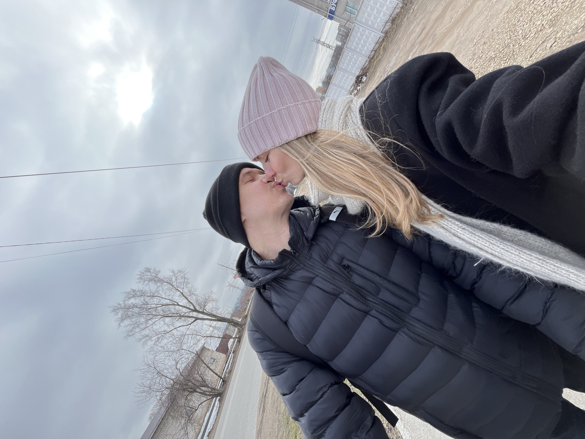 A couple, a man and a woman, are kissing outdoors on an overcast day. The man is wearing a black beanie and a black puffer jacket. The woman has long blonde hair and is wearing a light pink beanie, a white sweater, and a black coat. They are standing on a paved surface that appears to be a sidewalk or road. In the background, there is a building with Cyrillic writing on it, power lines, and bare trees. The setting is in the city of Chaikovsky, Russia.
