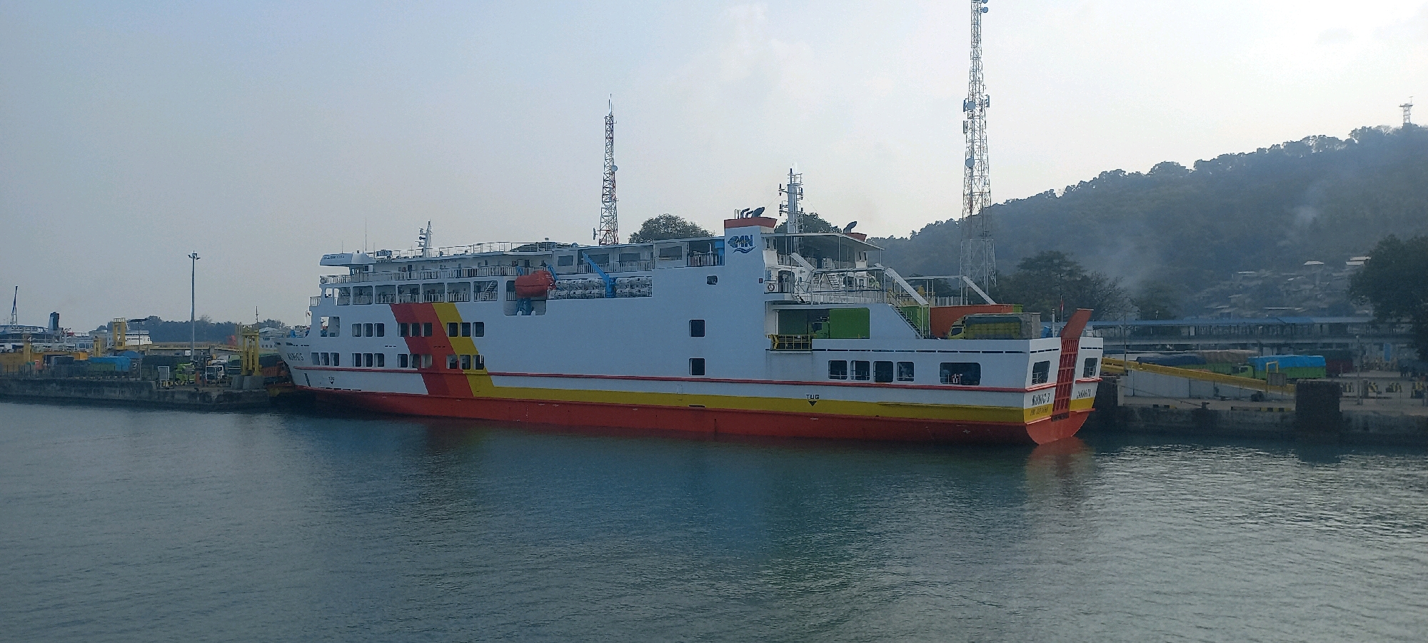 The image depicts a large passenger and vehicle ferry docked at a harbor, likely in Cilegon, Indonesia, given the presence of industrial structures and a hilly, vegetated background characteristic of the region. The ferry, predominantly white with red and yellow accents along its hull and upper decks, is a multi-story vessel with numerous windows. Several vehicles, including trucks, are visible on the dock behind the ferry, suggesting it is loading or unloading. Two prominent telecommunication towers stand tall in the background, situated on a densely wooded hillside. The sky is overcast with a light haze, and the water in the foreground is calm with a rippled surface. Several cranes and other port infrastructure are visible along the shoreline, indicating an active industrial port. The overall scene conveys a sense of maritime commerce and transportation in an Indonesian coastal city.