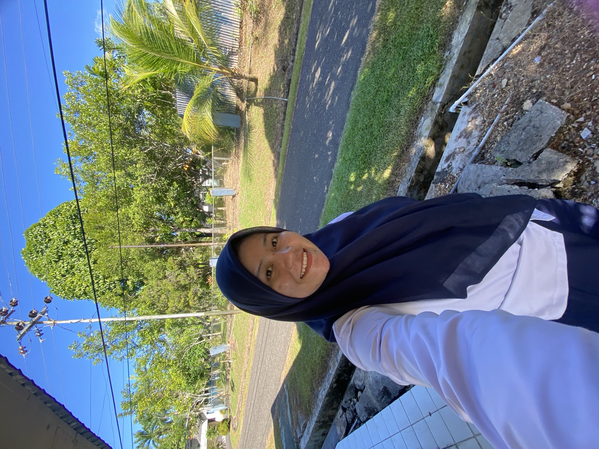 An Indonesian woman wearing a dark blue hijab and a white shirt is taking a selfie outdoors. She is smiling at the camera. The background shows a paved road, green grass, trees, and a utility pole with power lines. The scene appears to be in a residential or semi-rural area, possibly in the city of Banjarbaru, Indonesia, given the tropical vegetation and clear blue sky. The woman is positioned in the foreground, with the environment filling the rest of the frame. The lighting suggests it is daytime, likely morning or afternoon due to the strong sunlight and shadows.