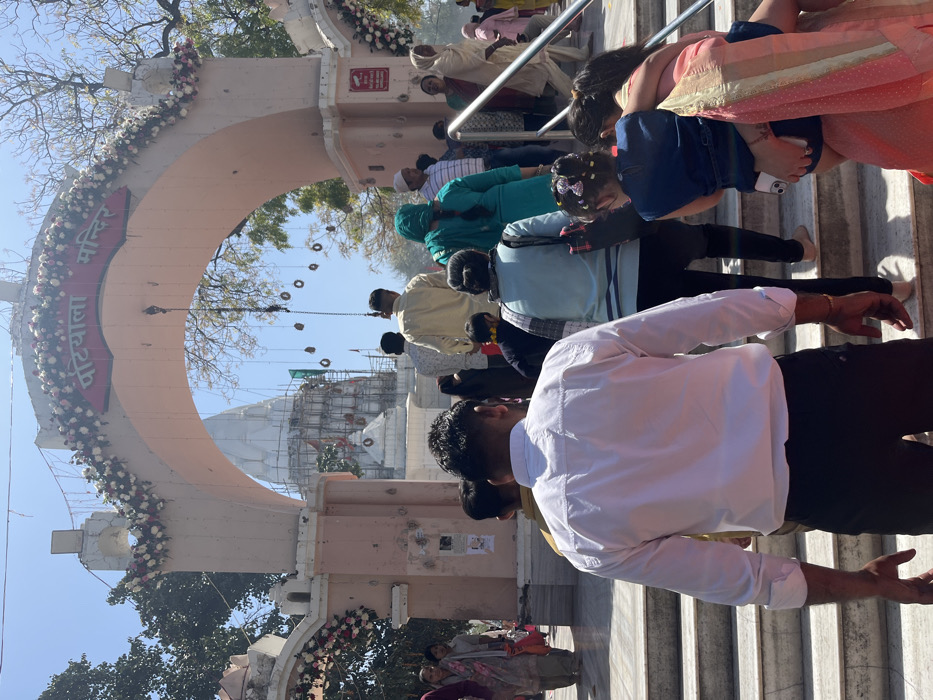 The image shows a group of people ascending a set of stairs leading to a temple entrance in Panchkula, India. The entrance is an archway adorned with flowers and the words "Patiyala Mandir" in Hindi. The temple, visible through the archway, is under construction. People are walking in different directions, with some holding hands and interacting with each other. One man, wearing a white shirt, is in focus and is seen ascending the stairs. A woman in a blue shirt is holding a young girl in a blue dress, who is also on the stairs. There are other people walking in the background. The weather is sunny and clear. The location, based on the coordinates provided, is near the Patiala Mandir temple on Sector 4, Panchkula.