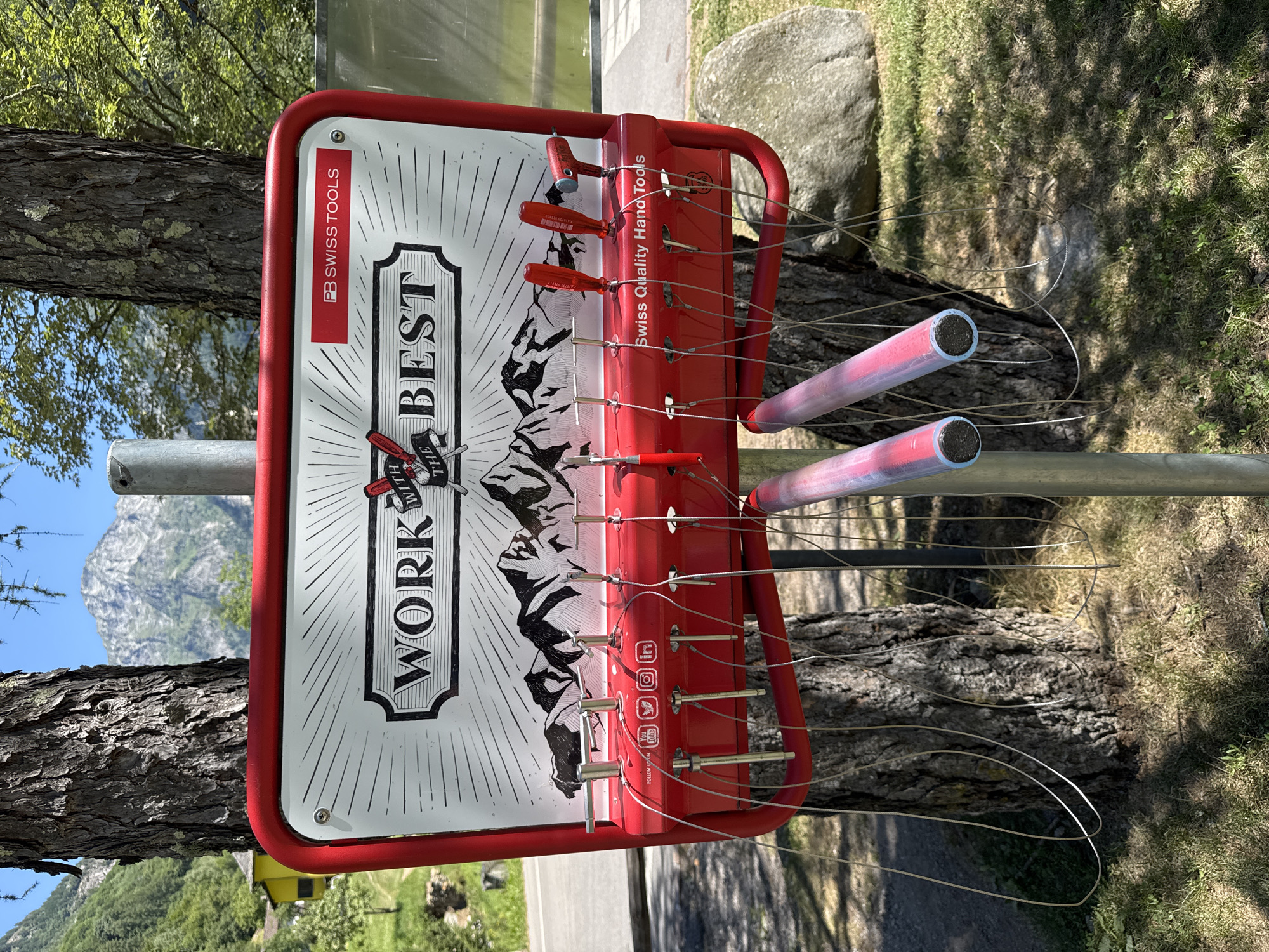 This image captures an outdoor bicycle repair station in Verzasca, Switzerland. The station is mounted on a metal pole positioned between two trees. The central object is a red metal frame holding a white sign that reads "WORK WITH THE BEST PB SWISS TOOLS." Below the sign, a red panel displays various tools suspended by thin wires. These tools include screwdrivers and a tire pump attachment. The background reveals a mountainous landscape, with a road and grassy areas visible. The station appears to be in a park or natural setting, indicated by the trees, rocks, and greenery. There are no people present in the image.