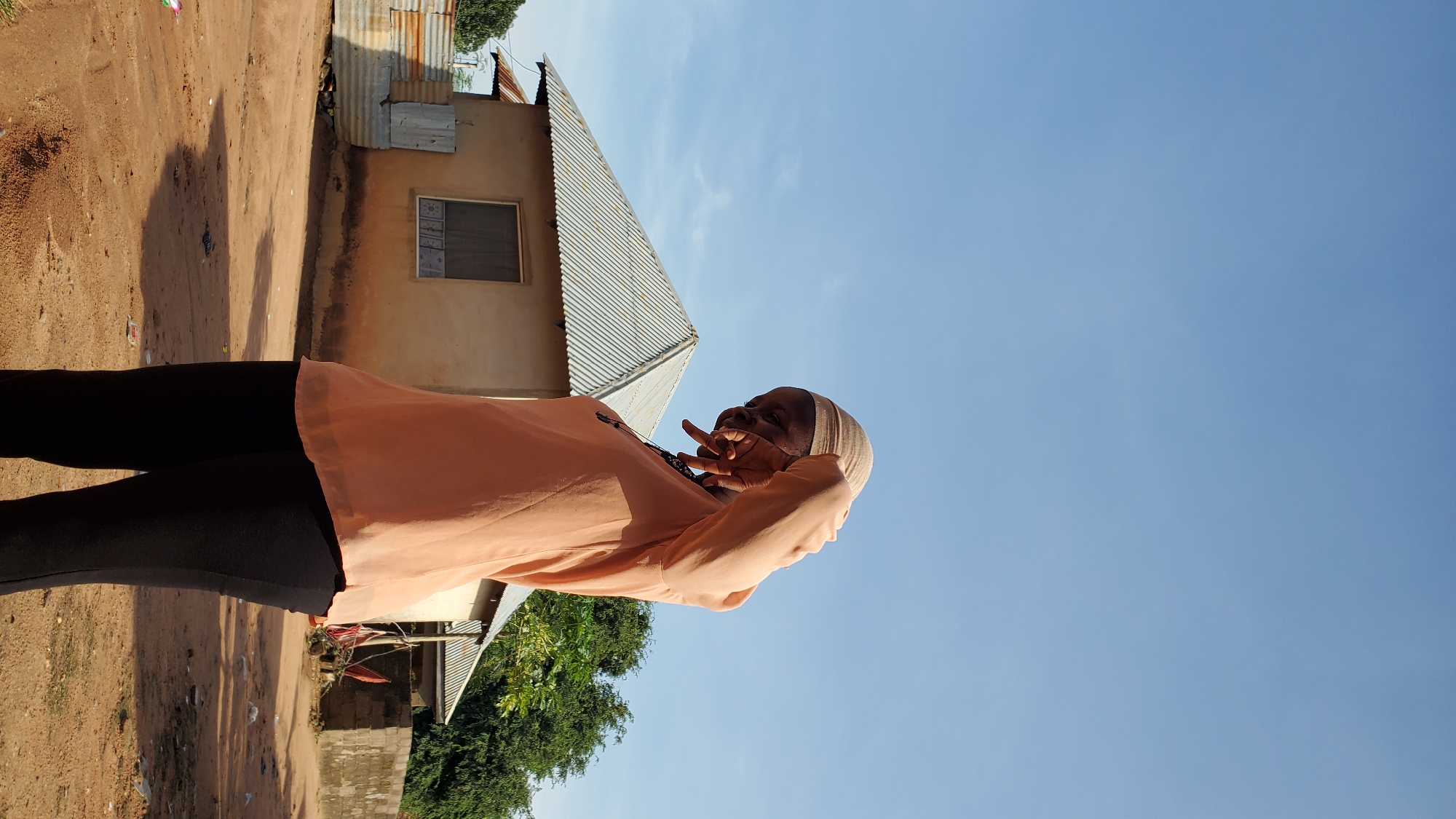 In the city of Jalingo, Nigeria, a young woman stands outdoors in a sandy, rural area. She is wearing a light peach-colored, sheer long-sleeved shirt and black leggings. Her head is covered with a beige hijab. She is looking down and to her left, with her right arm raised, obscuring part of her face as if shielding her eyes or wiping away a tear. Behind her, a modest, light-brown house with a corrugated metal roof is visible, along with some trees and other simple structures. The ground is dusty and uneven, with scattered debris. The sky above is clear and blue.