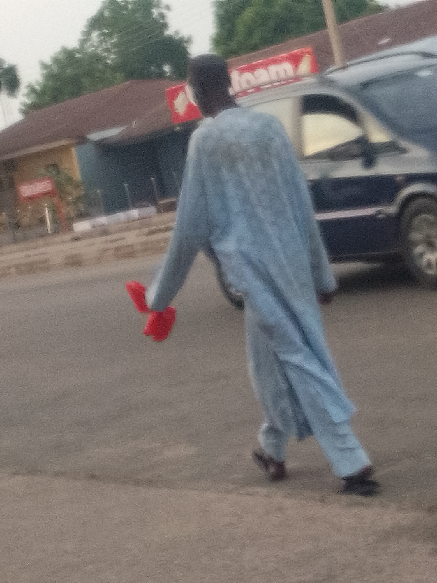 A person in a light blue garment walks away from the camera on a paved road in Pantame, Nigeria. They are holding a bright red object in their left hand, which appears to be a cloth or fabric. The person is wearing dark shoes. In the background, a dark-colored van is parked, and beyond that, a building with a reddish-brown roof is visible, along with some trees. A sign with white lettering on a red background is mounted on the building. The overall lighting suggests it might be late afternoon or early morning.