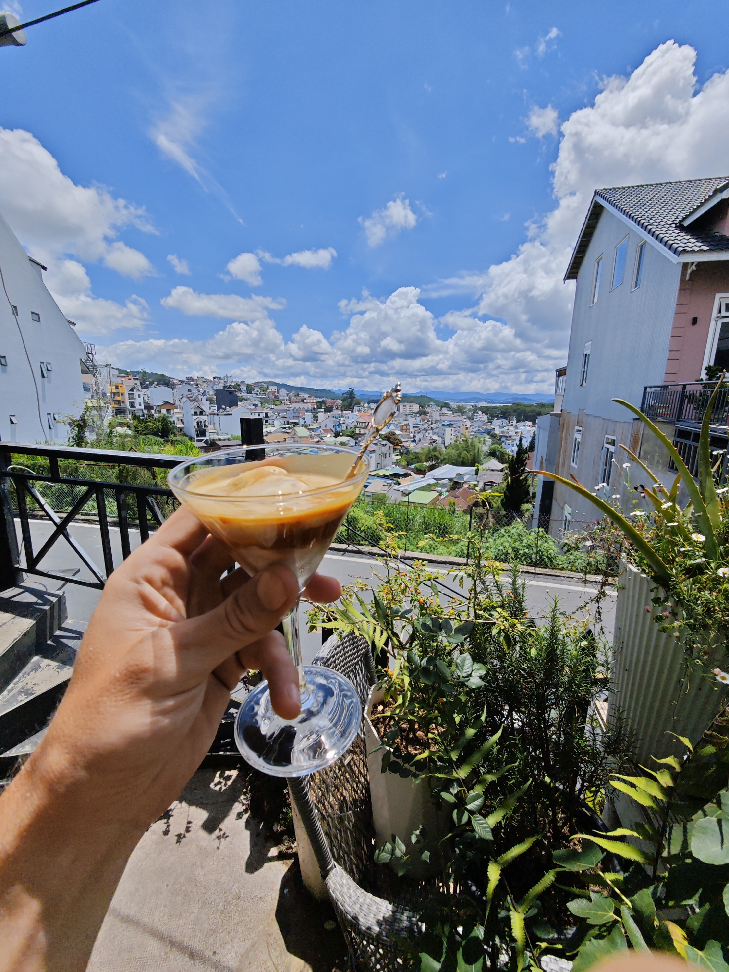 A hand holds a martini glass filled with an iced coffee drink, possibly an affogato, in the foreground. The drink has layers of dark coffee and lighter cream or ice cream. The hand and glass are positioned in front of a scenic view of a town nestled in a valley, with buildings of various sizes and colors spread across the landscape. In the background, hills and mountains stretch towards the horizon under a bright blue sky dotted with fluffy white clouds. To the right of the glass, lush green plants, including what appears to be an aloe vera plant with small white flowers, are growing in pots. A glimpse of a grey building with a dark tiled roof is visible on the right side of the image. On the left, a white building with visible windows and an exterior staircase is partially visible. The scene appears to be taking place outdoors on a balcony or patio, suggesting a pleasant day. The overall mood is relaxed and scenic, hinting at a vacation or leisurely moment. The location is identified as Da Lat, Vietnam. There is no visible text in the image.