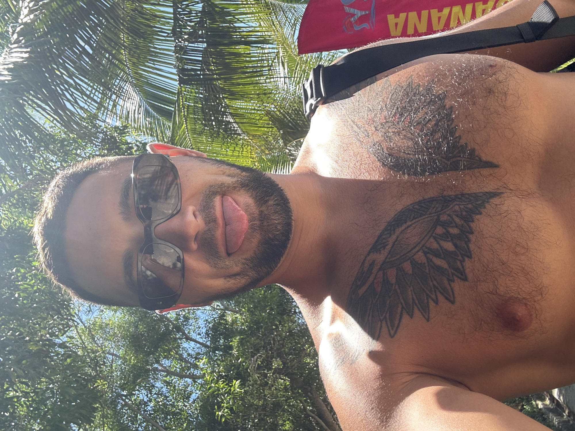A man is taking a selfie outdoors, with trees and palm fronds visible in the background. He is wearing sunglasses and has his tongue sticking out. He has tattoos on his chest. A red flag with the word "BANANA" is partially visible on the right side of the frame. The lighting suggests it is a sunny day. The location is not specified as Bac Nha Trang Ward, Vietnam, as there are no visual cues to support this.