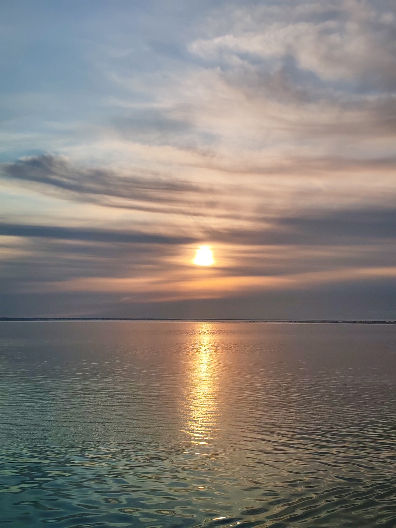 A serene sunset over the water is captured in this vertical, full-frame, eye-level shot. The sun, a bright, white orb, is positioned just above the horizon, casting a shimmering golden reflection across the rippling water. Wispy clouds, painted in hues of grey, orange, and blue, streak across the sky, creating a dramatic and tranquil atmosphere. The water itself is a mosaic of blues and greens, with the golden light of the sunset accentuating the gentle waves. The distant horizon is a thin, dark line separating the water from the sky, hinting at a vast, open expanse. The scene appears to be a natural landscape, possibly a bay or lake, with no visible signs of human activity or structures. The overall impression is one of peace and natural beauty. This scene is likely located in Veregonets, Russia, a region known for its picturesque bodies of water and stunning sunsets.